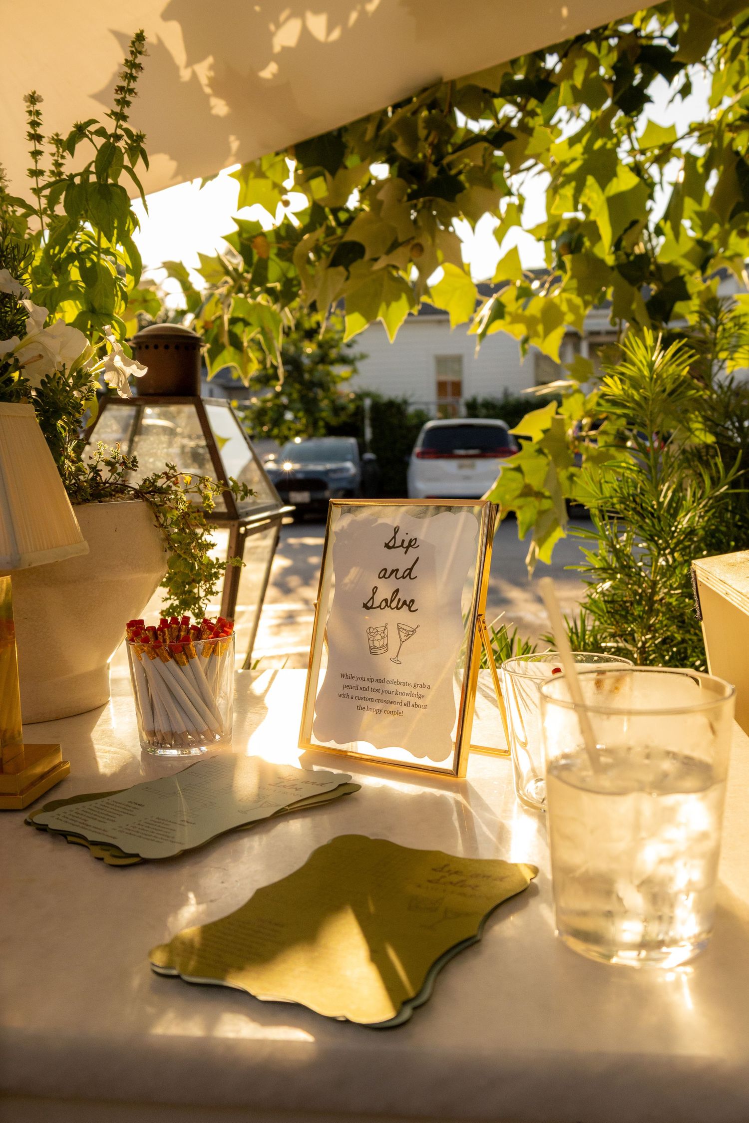 Outdoor table setting with matches, lantern, Sip and Solve sign, glassware, and green napkin under ivy in golden light.