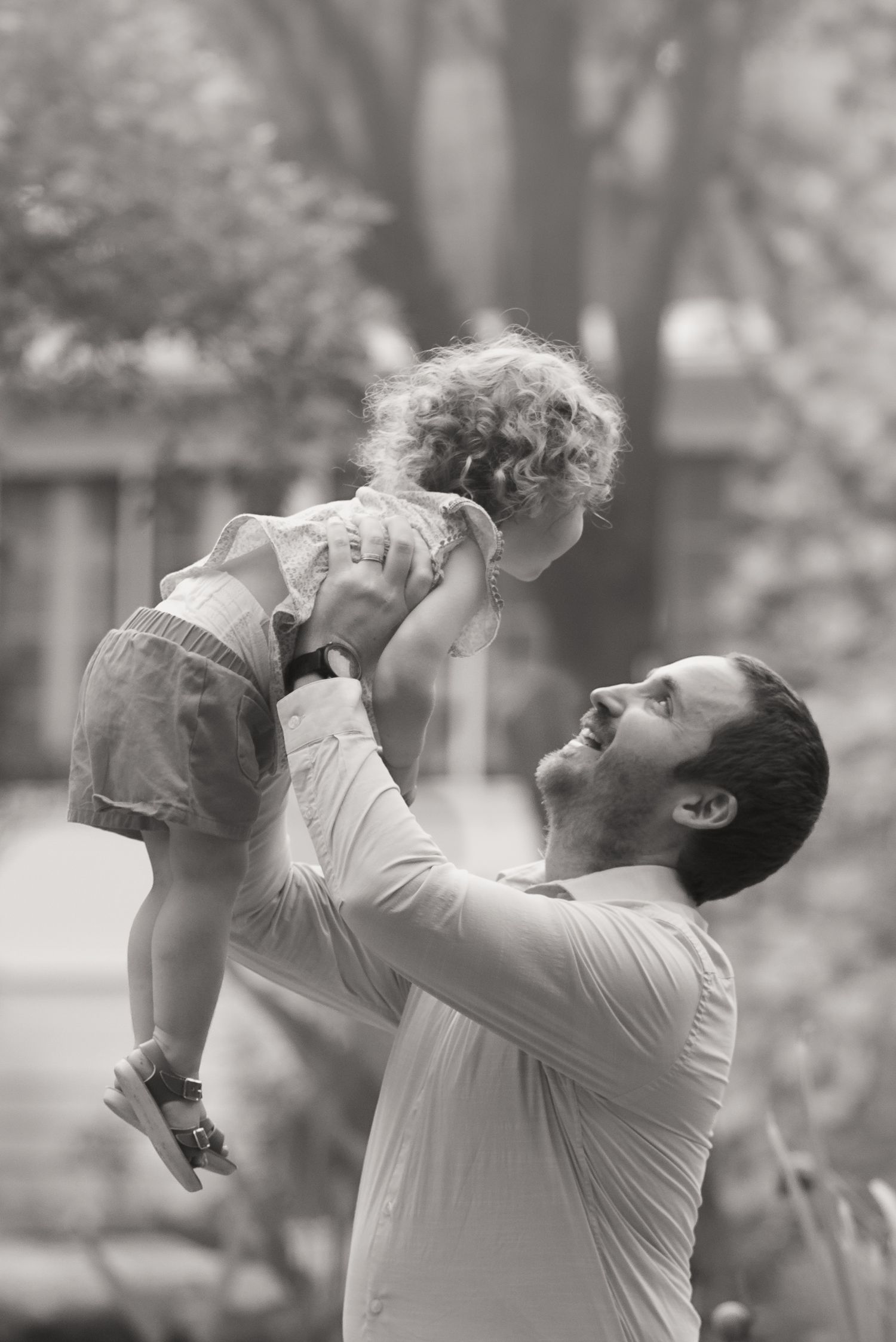 Black and white photo of a smiling father lifting his curly-haired toddler daughter in the air outdoors.