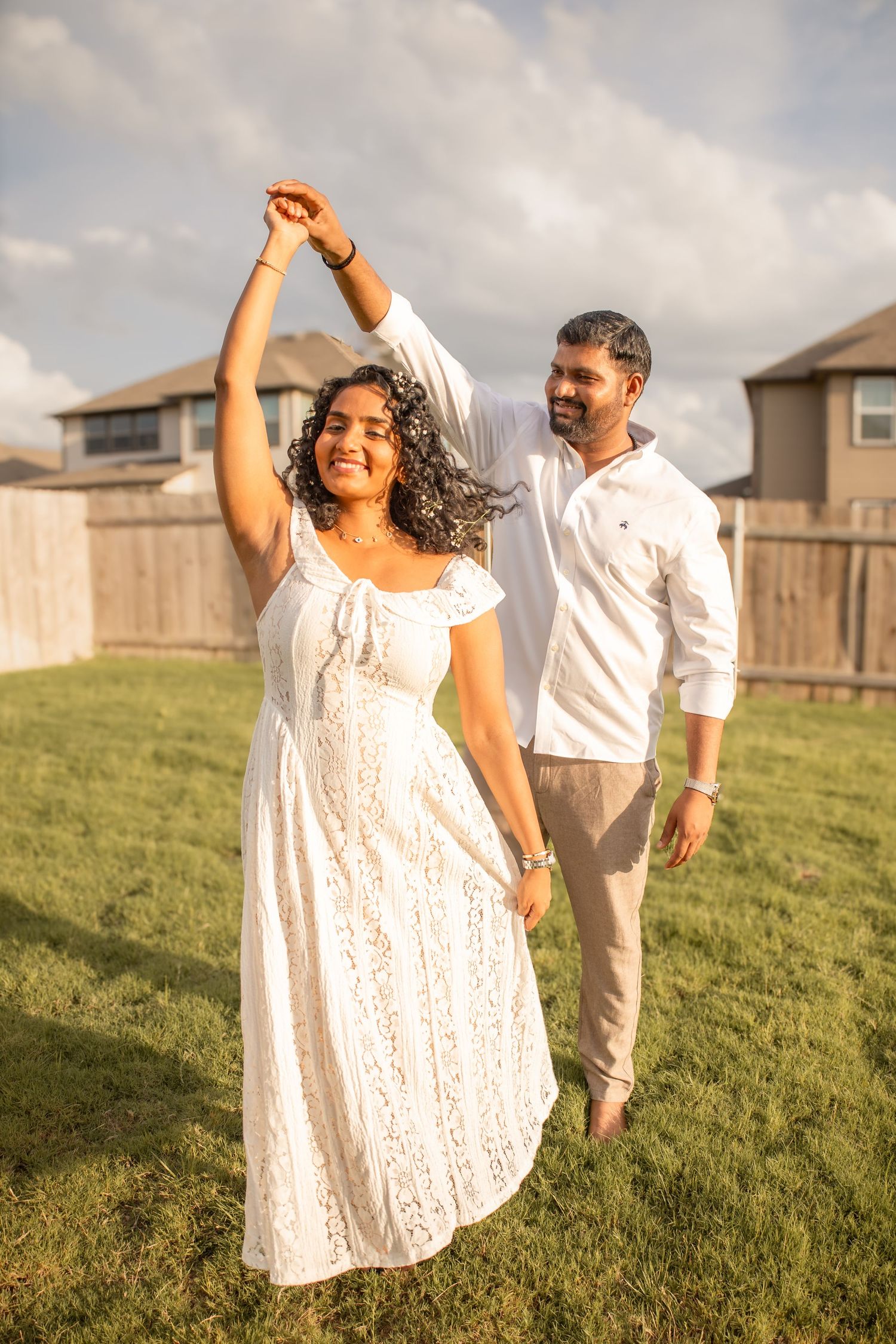 A joyful couple dances outdoors on green grass, woman in white lace dress being twirled by man in white shirt.