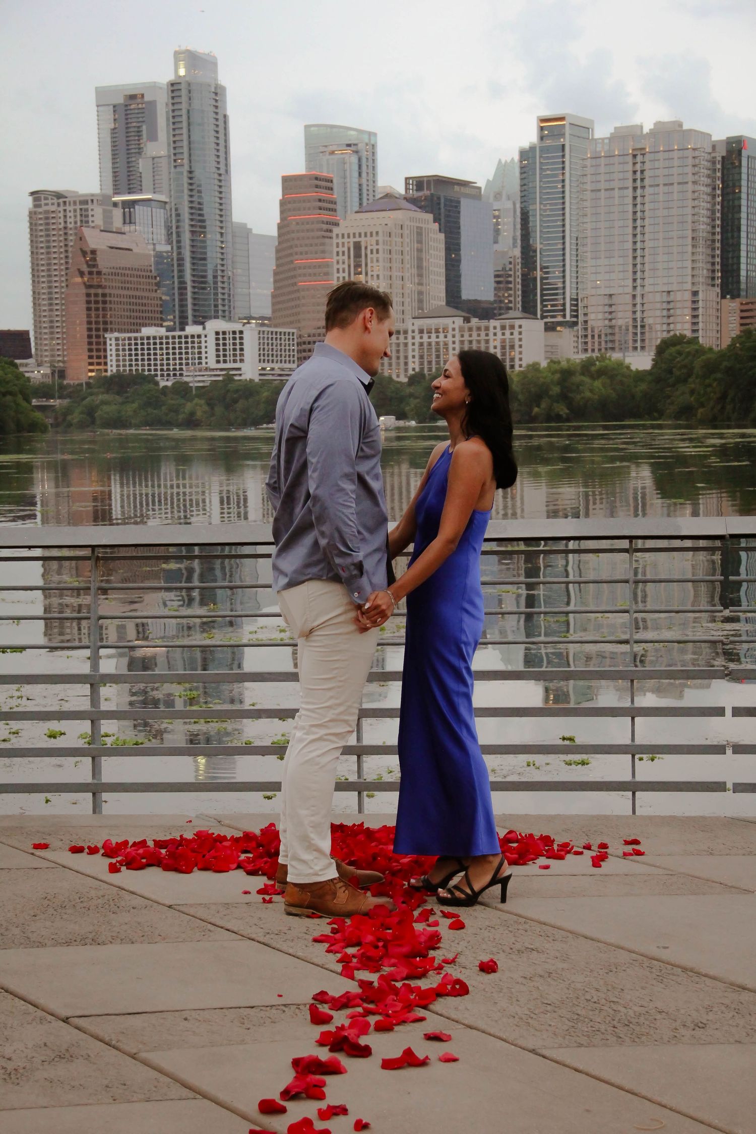 Newly engaged couple holding hands and smiling by a waterfront with a stunning city skyline view.
