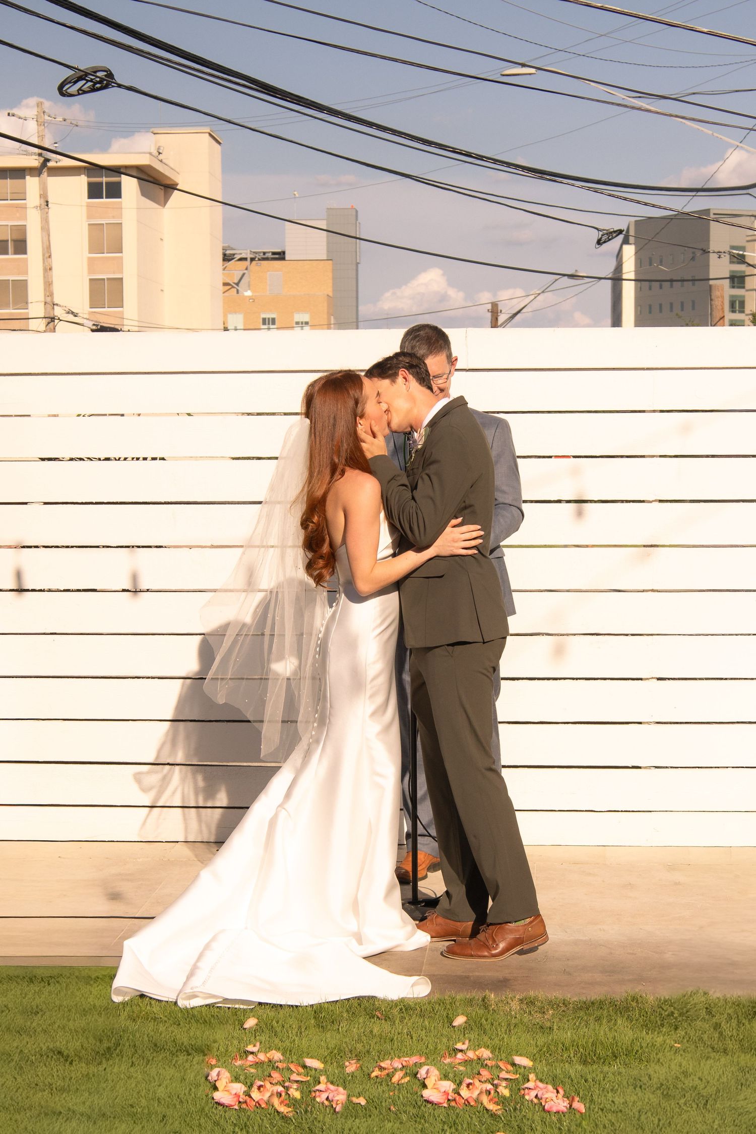 Bride and groom share a romantic kiss in front of a white fence at their outdoor wedding ceremony.