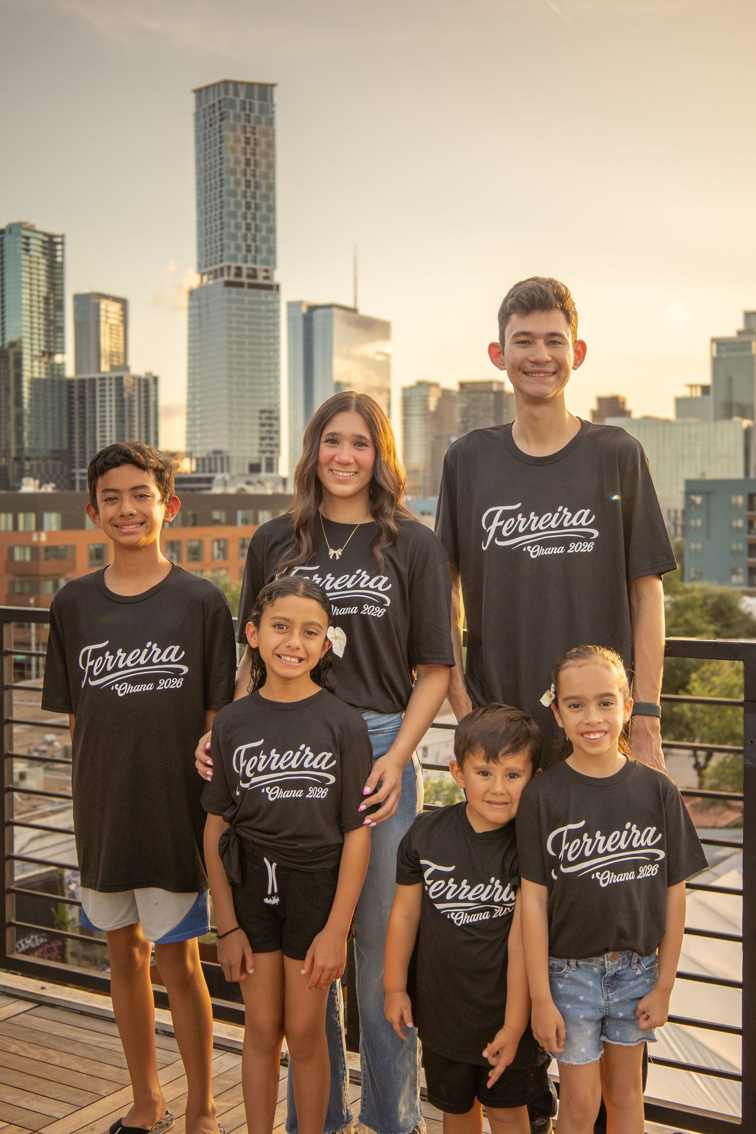 Six kids wearing matching black Ferreira family reunion shirts pose together on a rooftop with a city skyline at sunset.