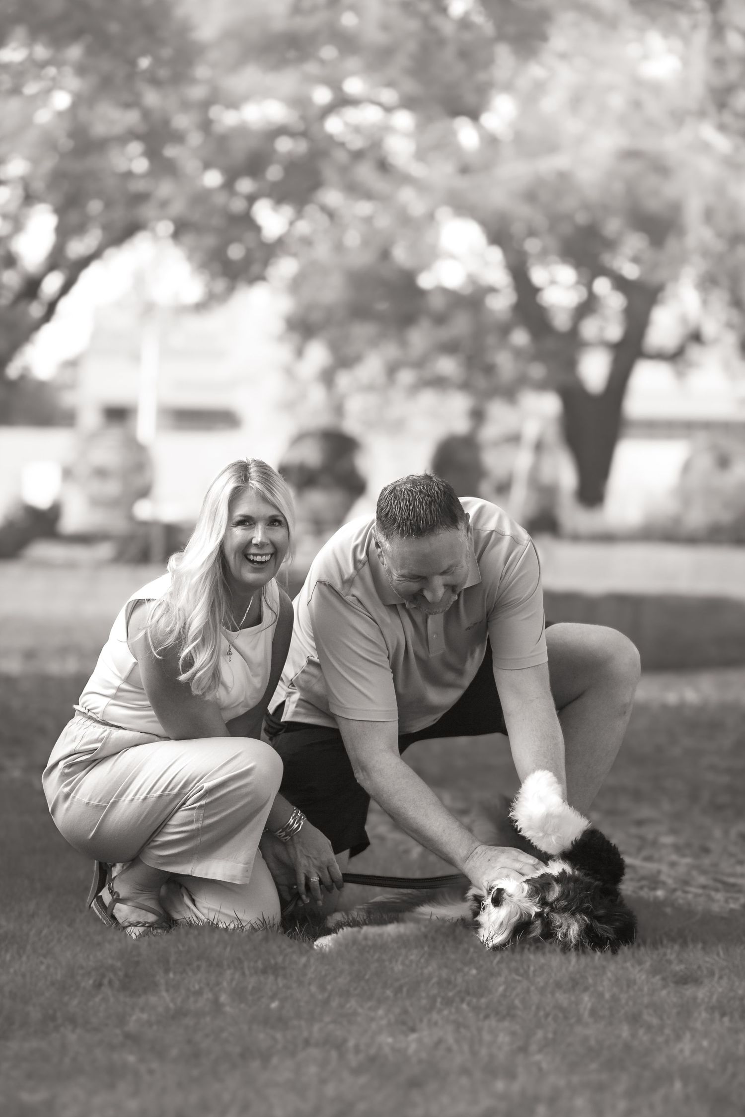 Couple crouching on grass playing with small dog in black and white park photo.
