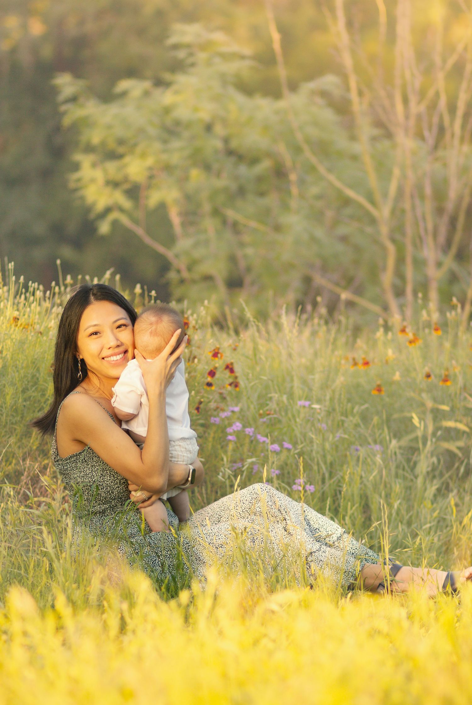 Smiling mother sitting in wildflower field holding baby close in warm golden sunlight.