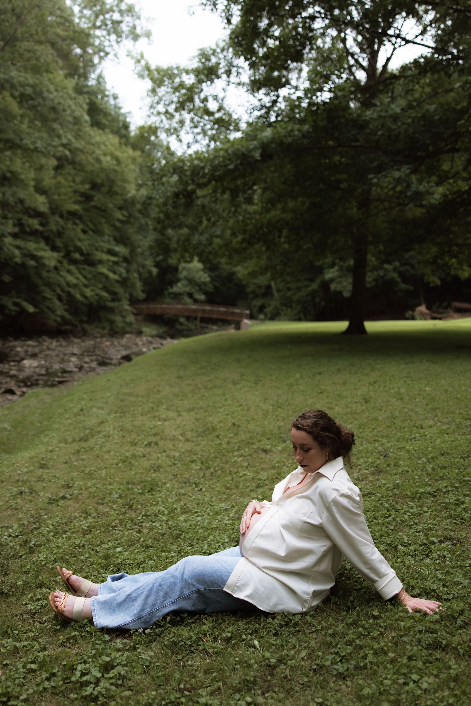 Pregnant woman in white shirt relaxing on grass near a tree-lined creek.