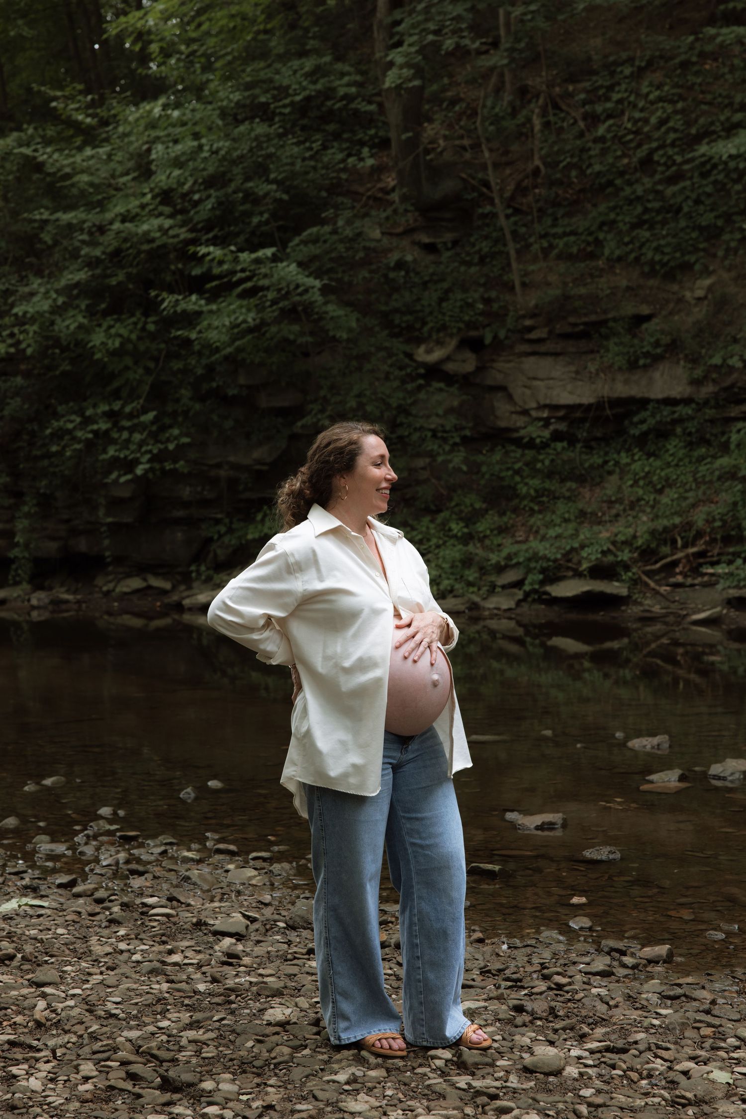Expectant mother in white shirt and jeans standing by a serene creek in a forest setting.