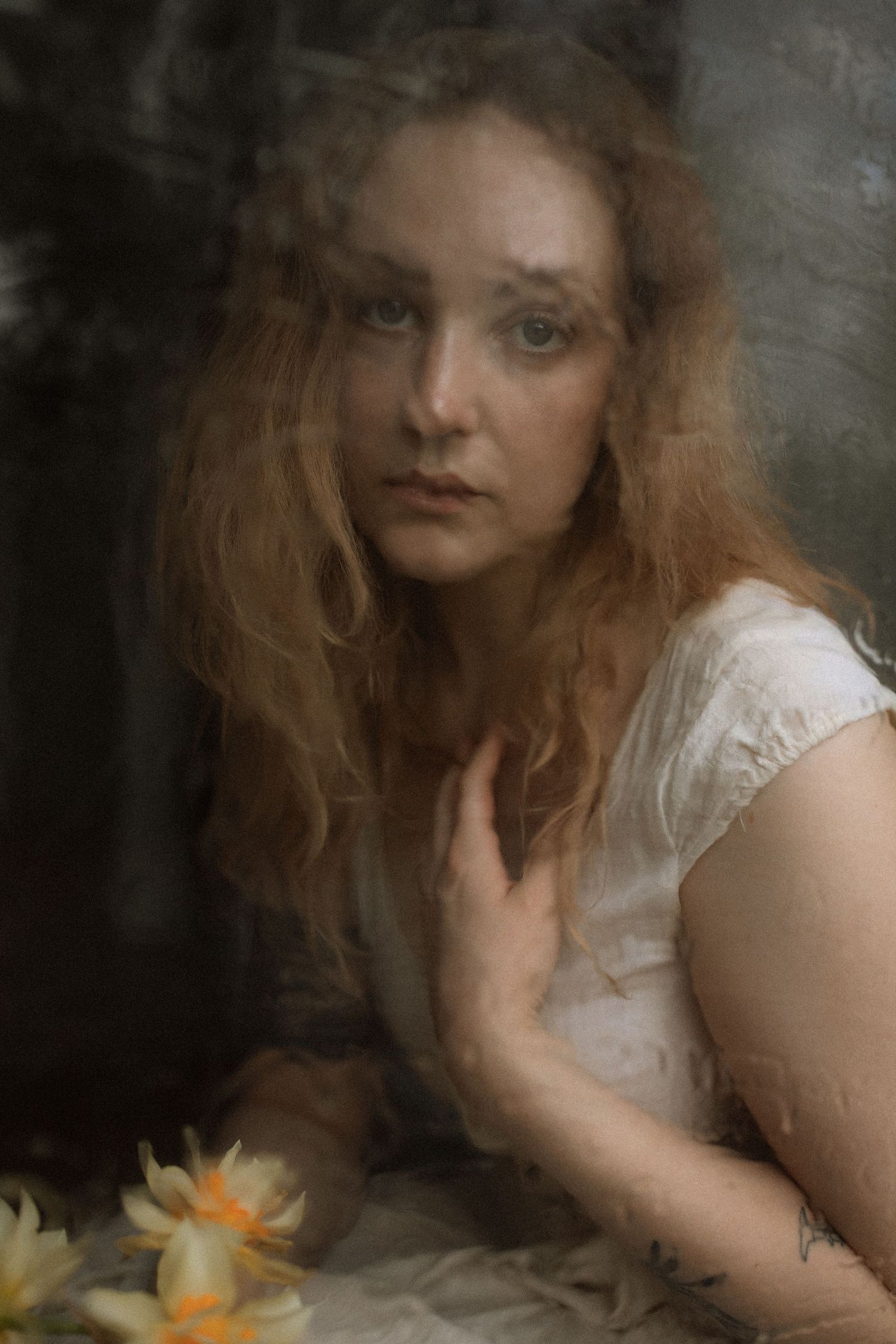 Ethereal portrait with flowing red hair in soft focus with daffodils, featuring white dress and moody dark background.