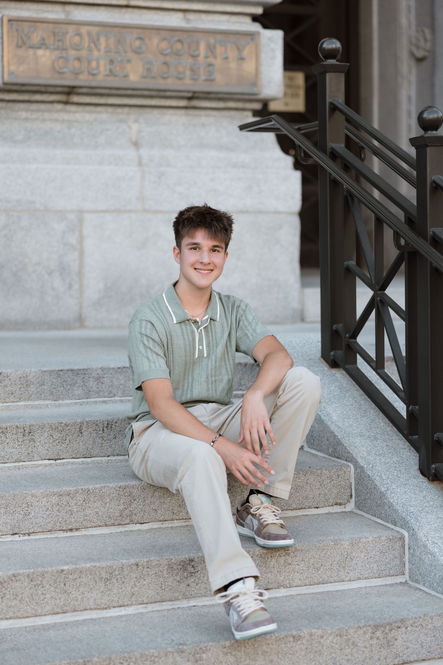 A young person in a striped shirt and khaki pants sits casually on concrete steps outside a building.