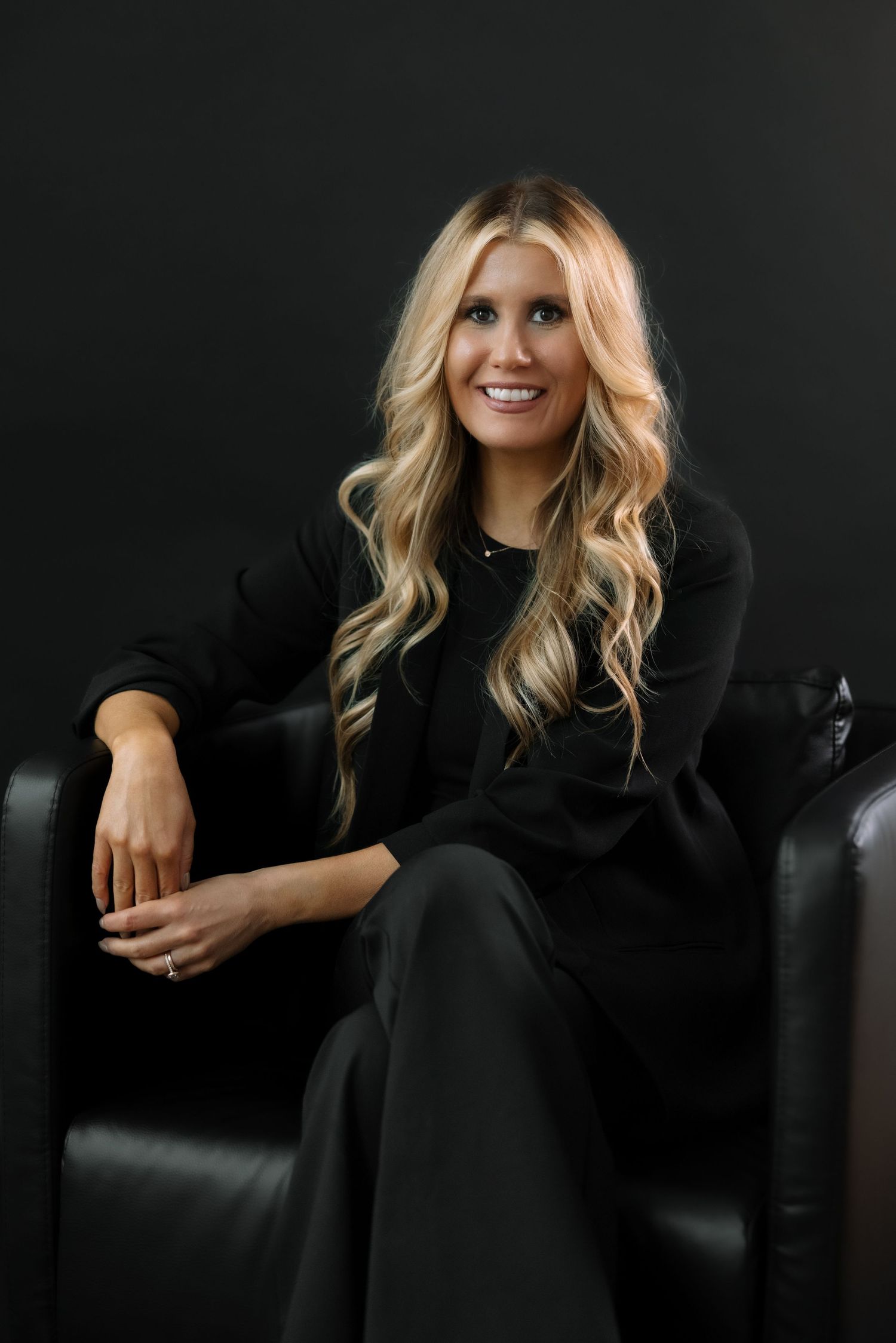 Professional studio portrait of a person with long blonde waves wearing black attire while sitting relaxed in a leather chair.