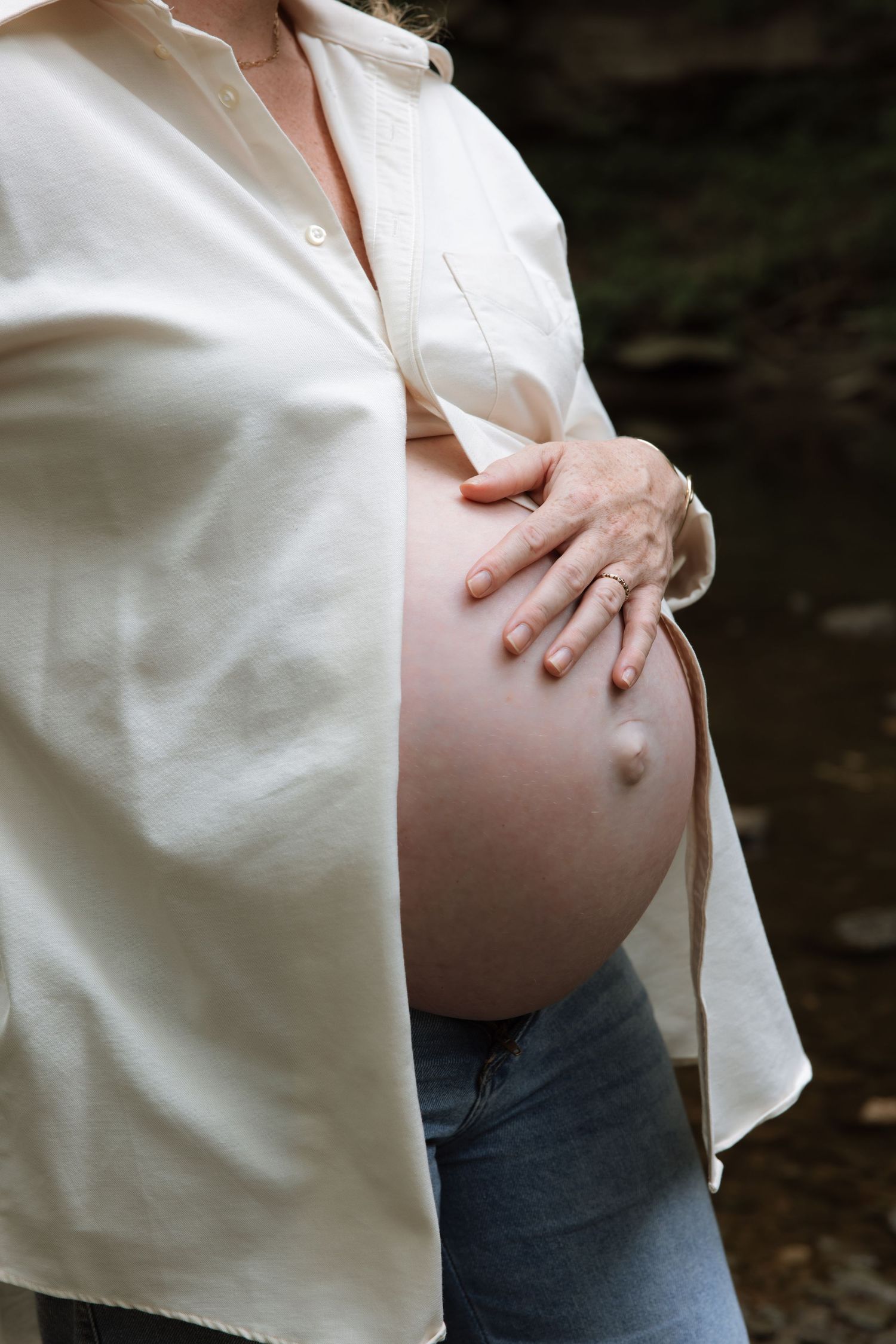 A pregnant person wearing a white button-up shirt and jeans tenderly holds their belly in a maternity photo.