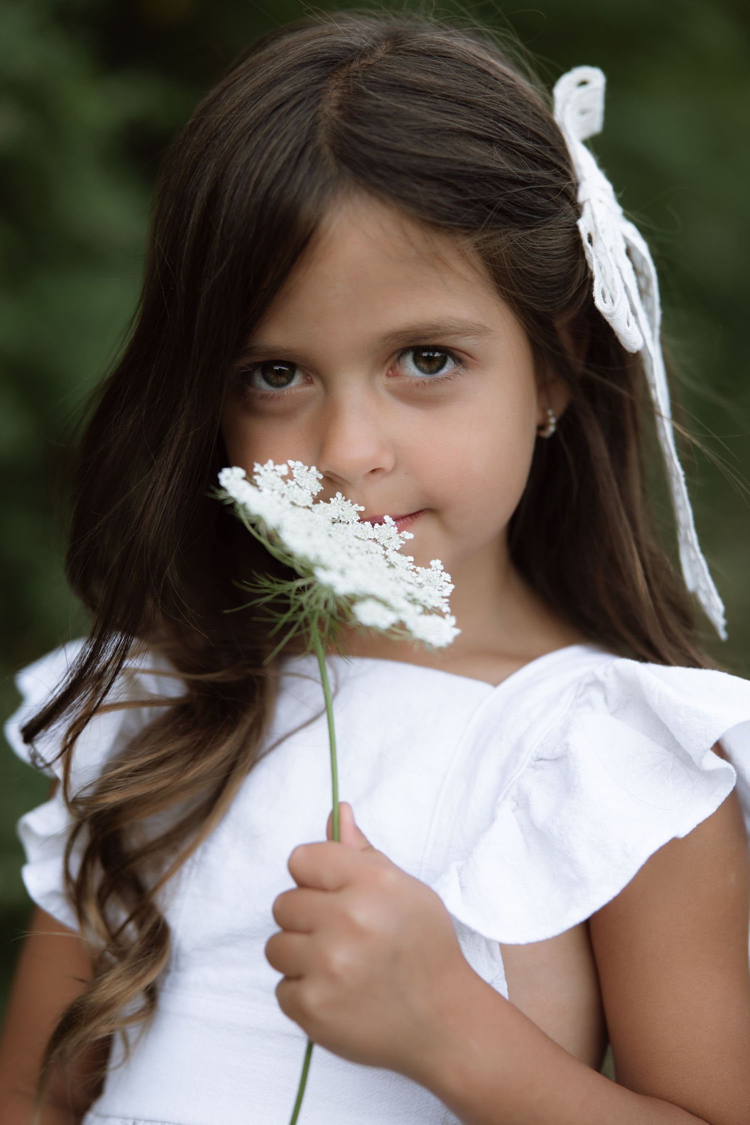 A child holding a delicate white flower up to their face while wearing a white dress outdoors.