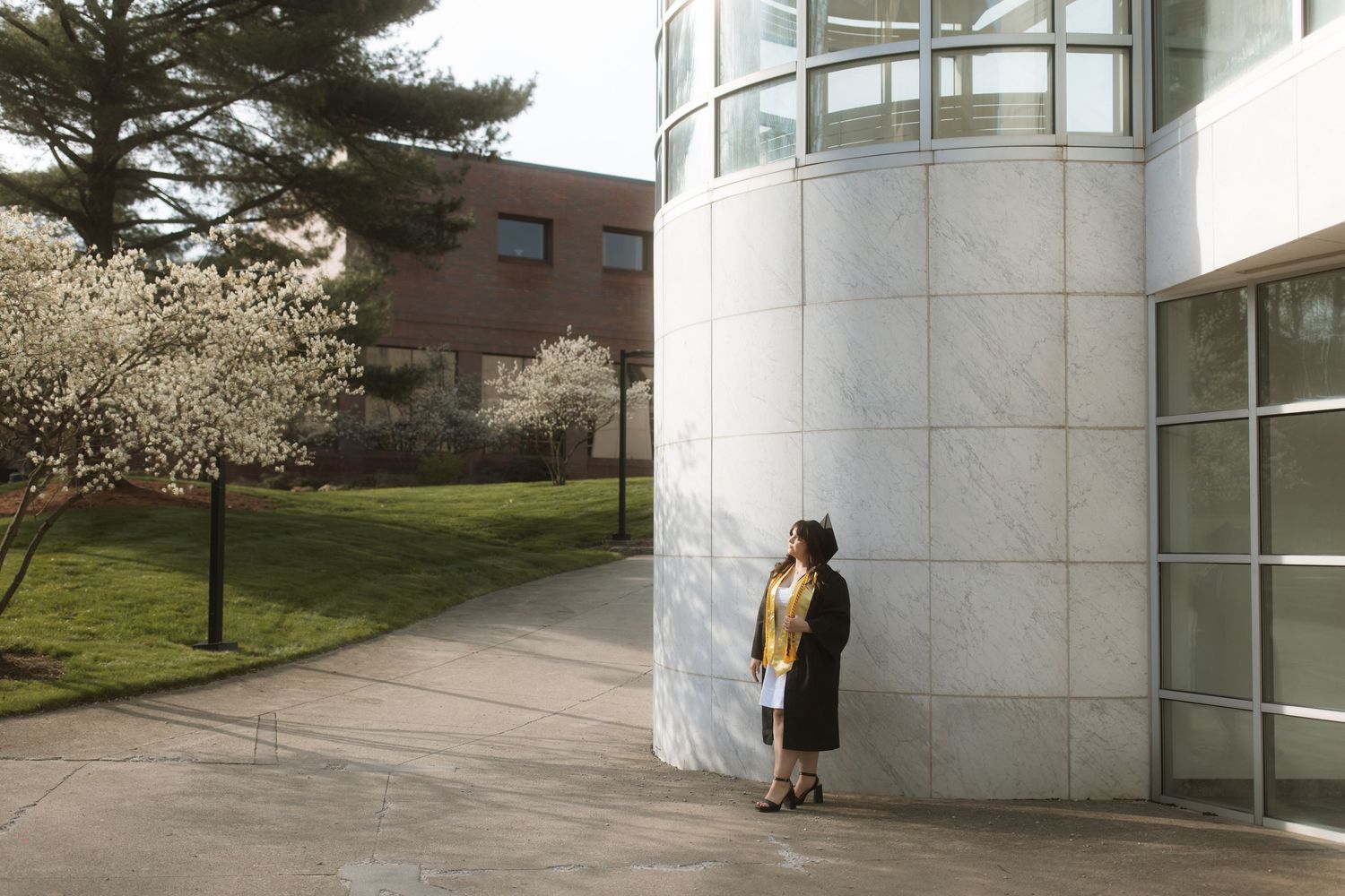 A person in yellow stands against a curved white building wall on a college campus with spring trees blooming nearby.