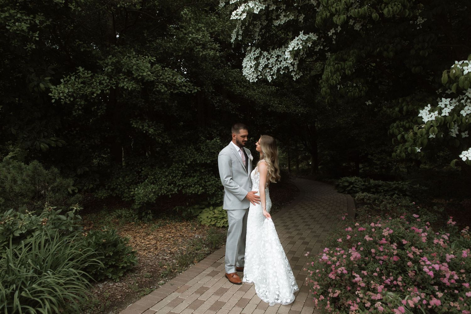 A romantic wedding portrait of a couple on a garden path surrounded by lush greenery and pink flowers.