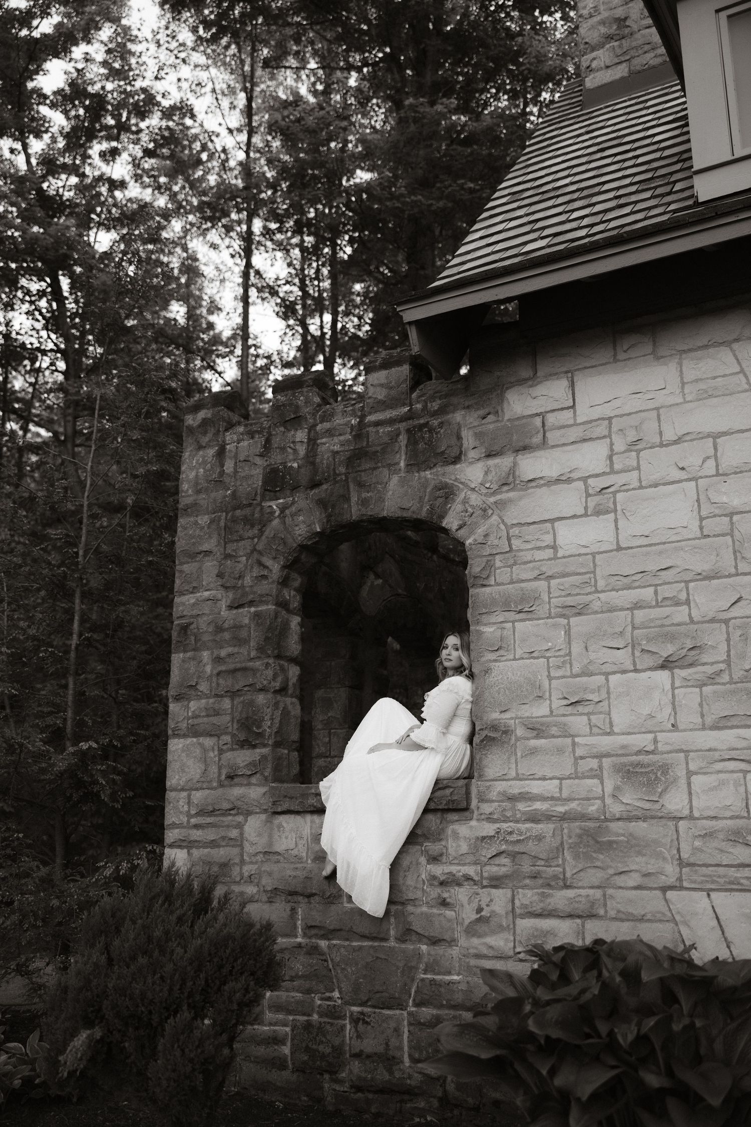 A vintage black and white photo shows a figure in white sitting in an arched stone window alcove of a rustic building.