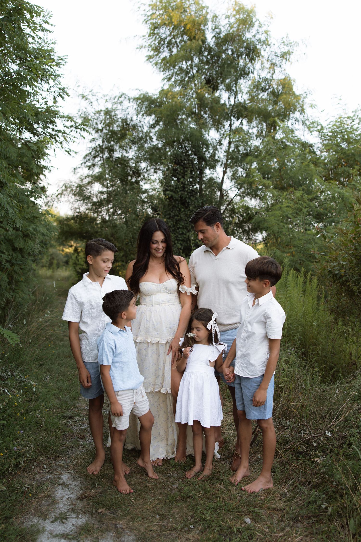 A family stands barefoot together outdoors wearing coordinating white and light blue outfits against lush greenery.