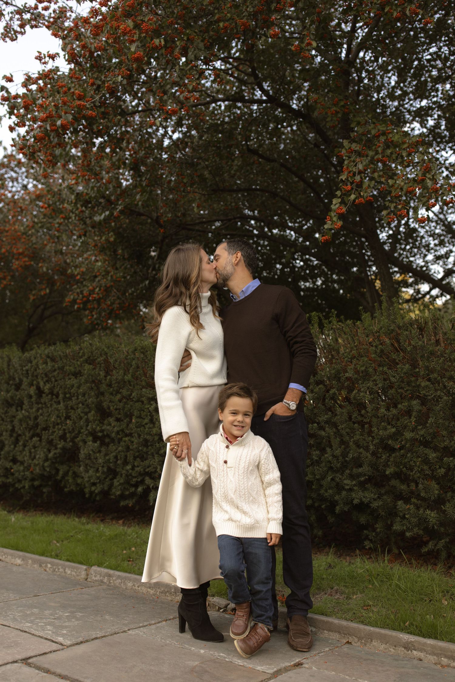A family in coordinating cream and brown outfits pose together outdoors during autumn with fall foliage in the background.