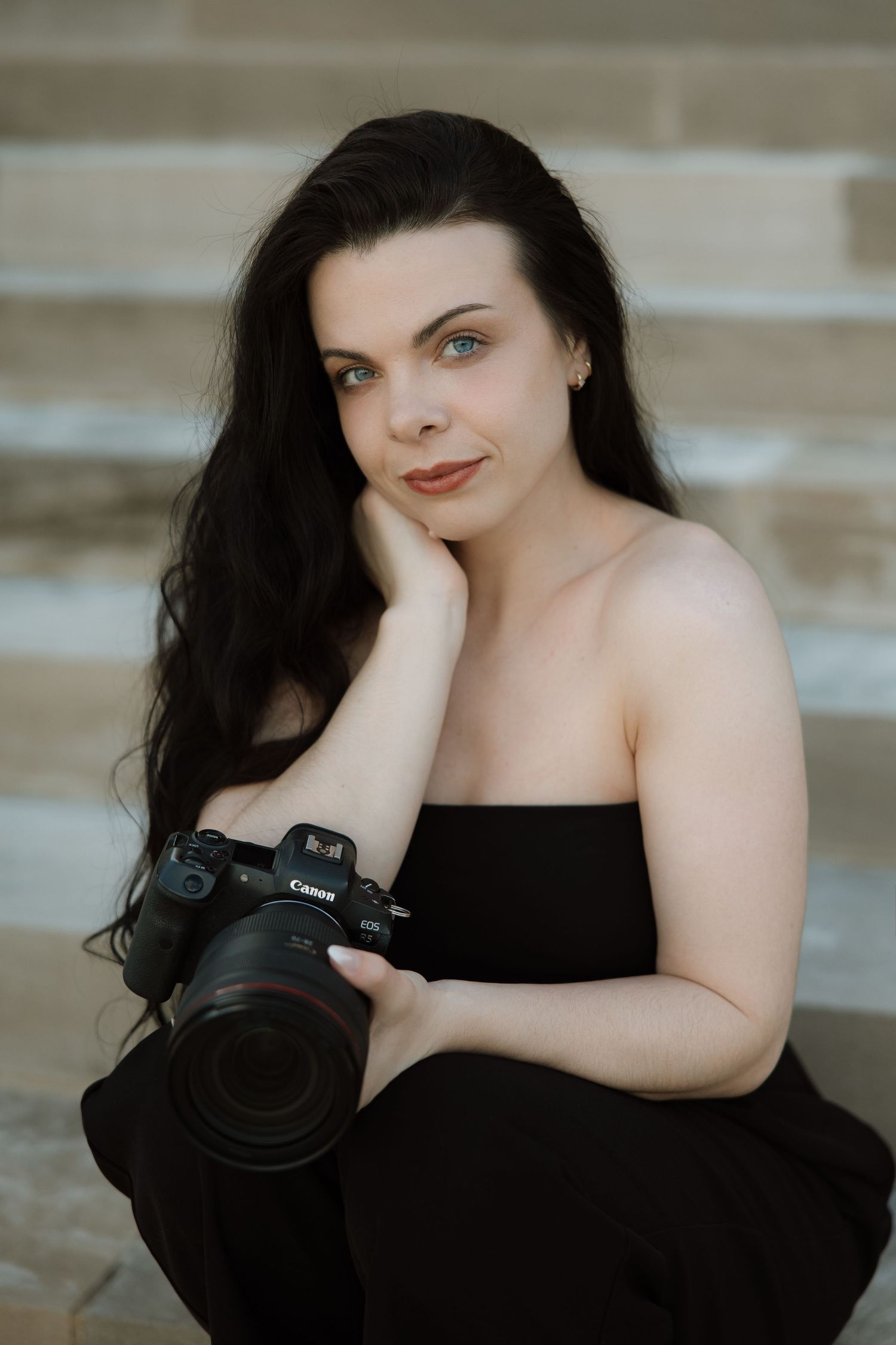 Woman with dark hair holding a Canon camera, sitting on stone steps in a black strapless outfit.