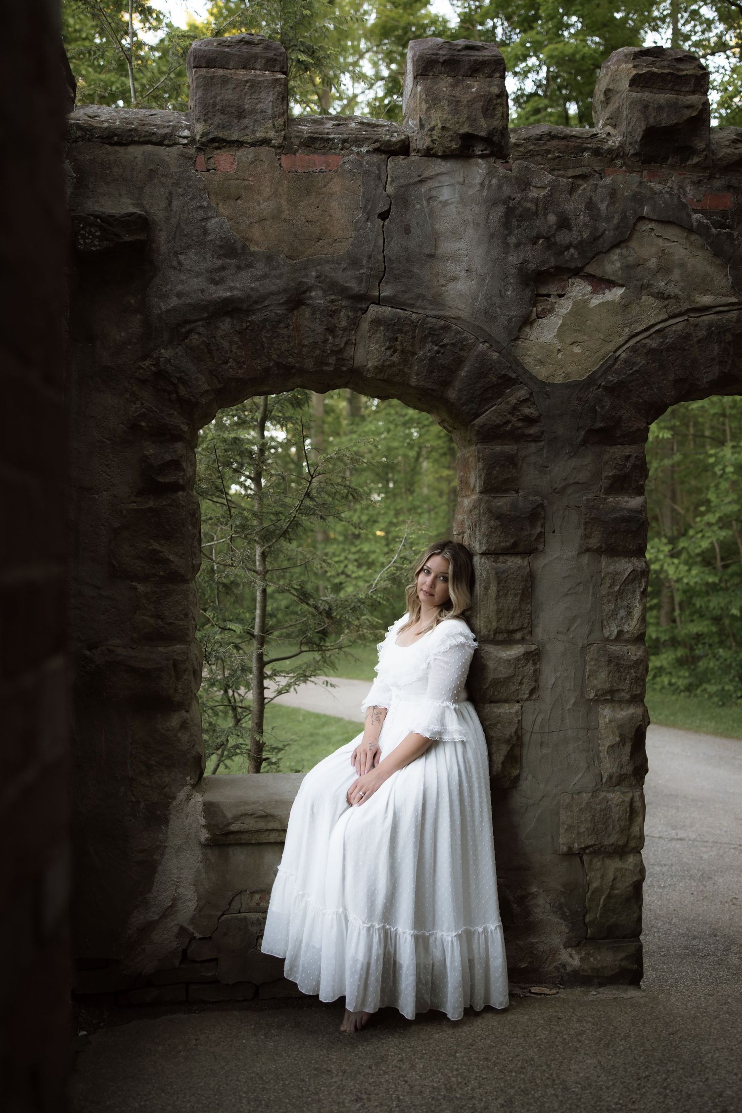A stone archway nestled in a wooded park provides a natural frame for someone in a flowing white dress at dusk.