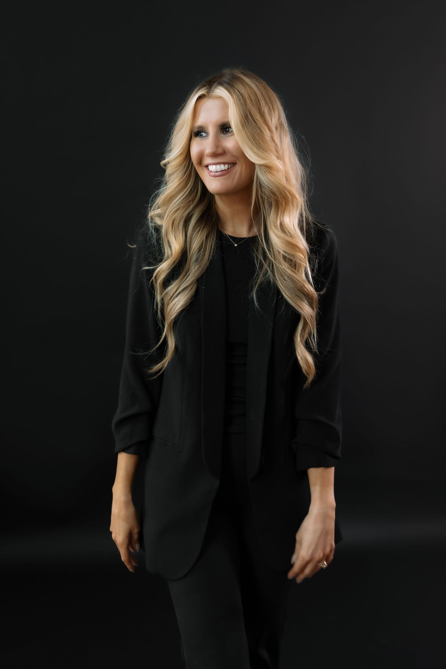 Professional studio portraits in black on black showing model wearing dark blazer and top with wavy blonde hair against dark backdrop.