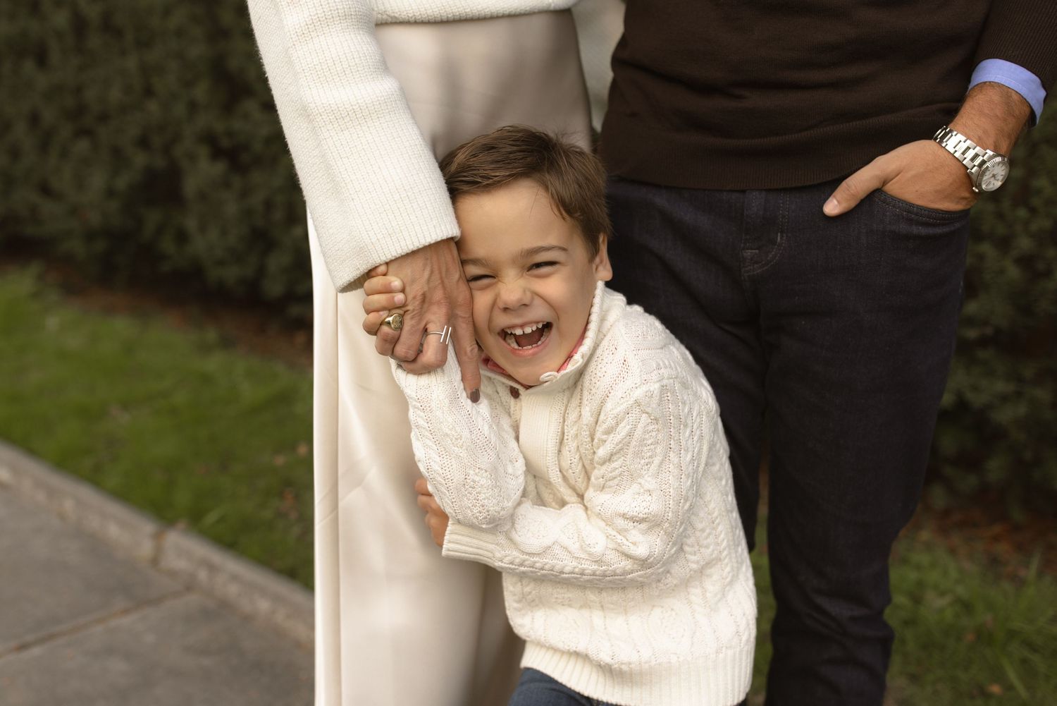 A small child in a white knit sweater leans against someone's legs while laughing joyfully outdoors.