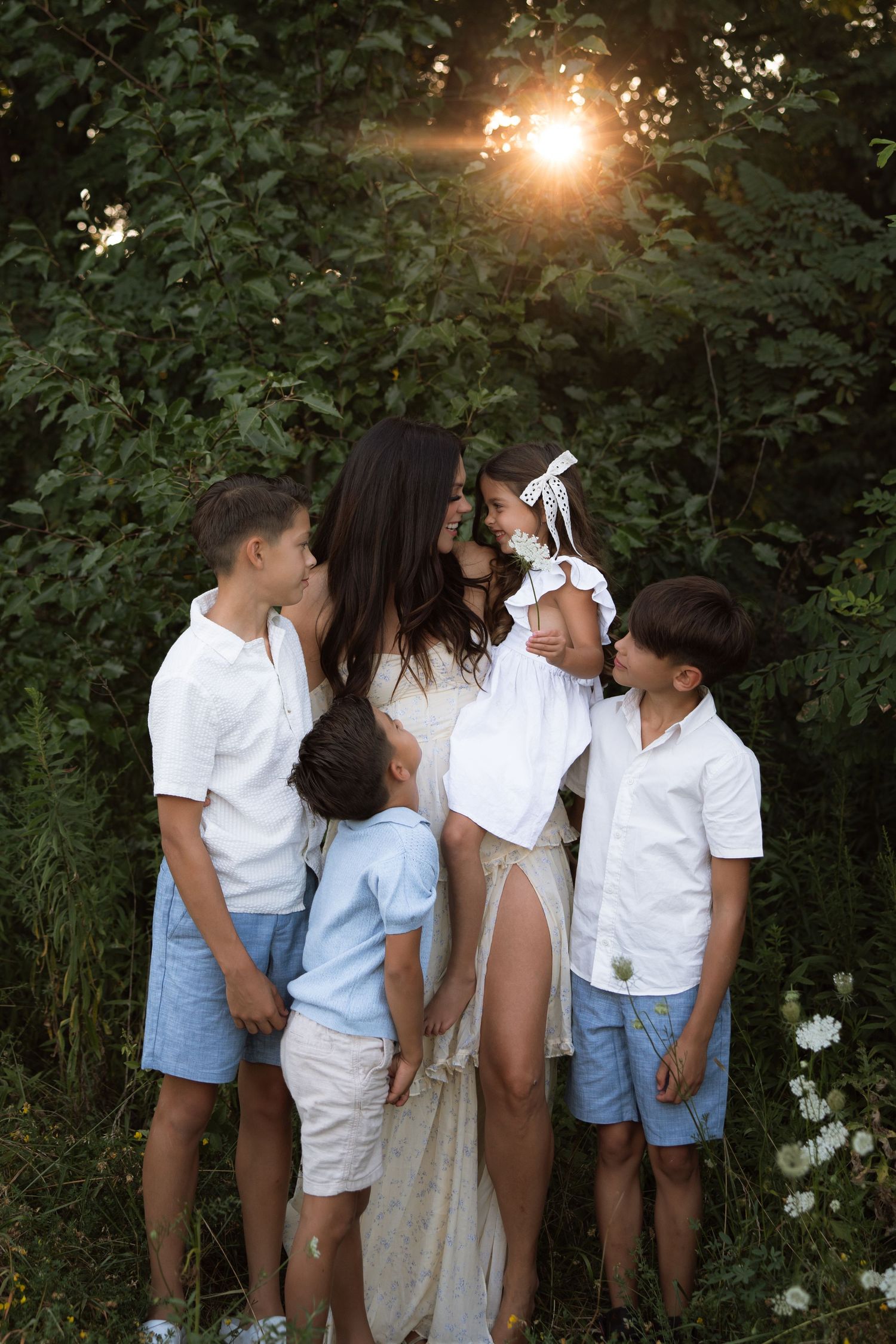A family wearing white and blue outfits poses together in a garden with sunlight filtering through trees at sunset.
