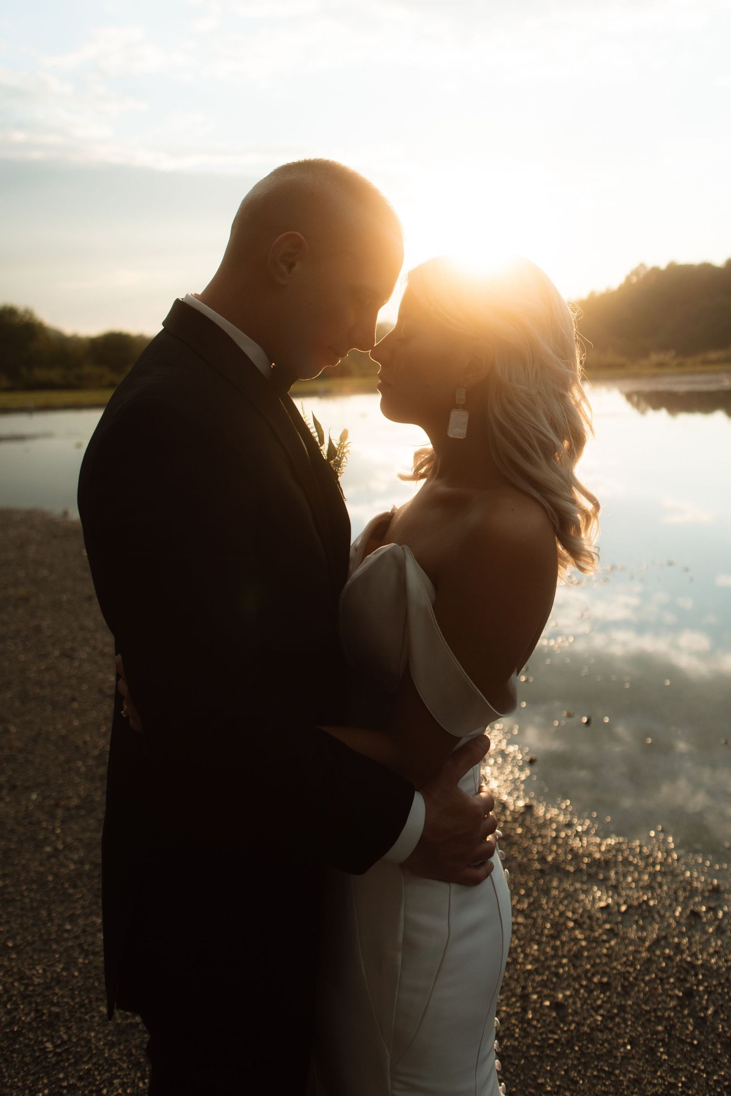 A couple embraces by a tranquil lake at sunset, silhouetted in romantic golden light as their figures reflect on the water.