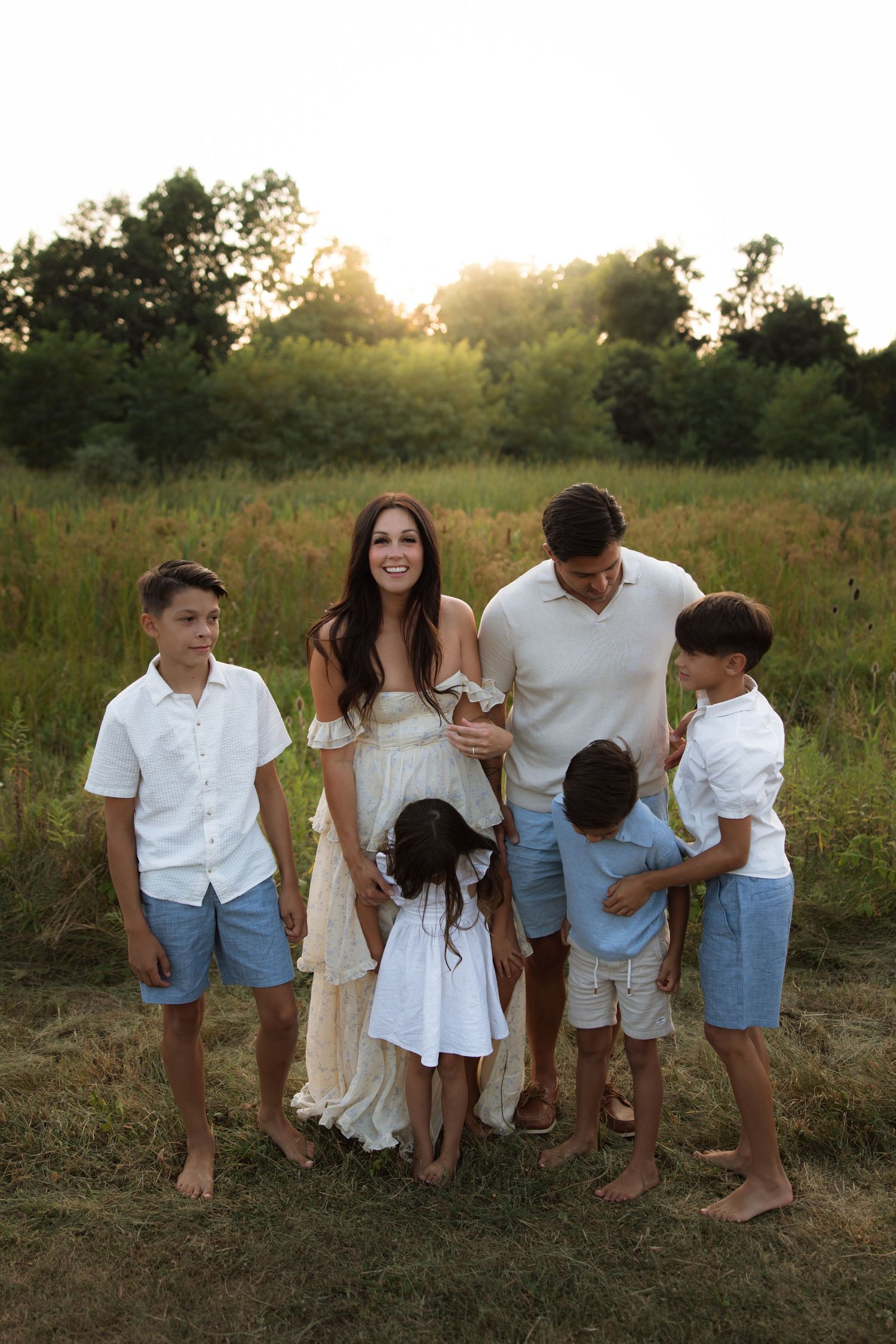 Family wearing coordinated white and blue outfits poses together in sunlit summer field at golden hour.