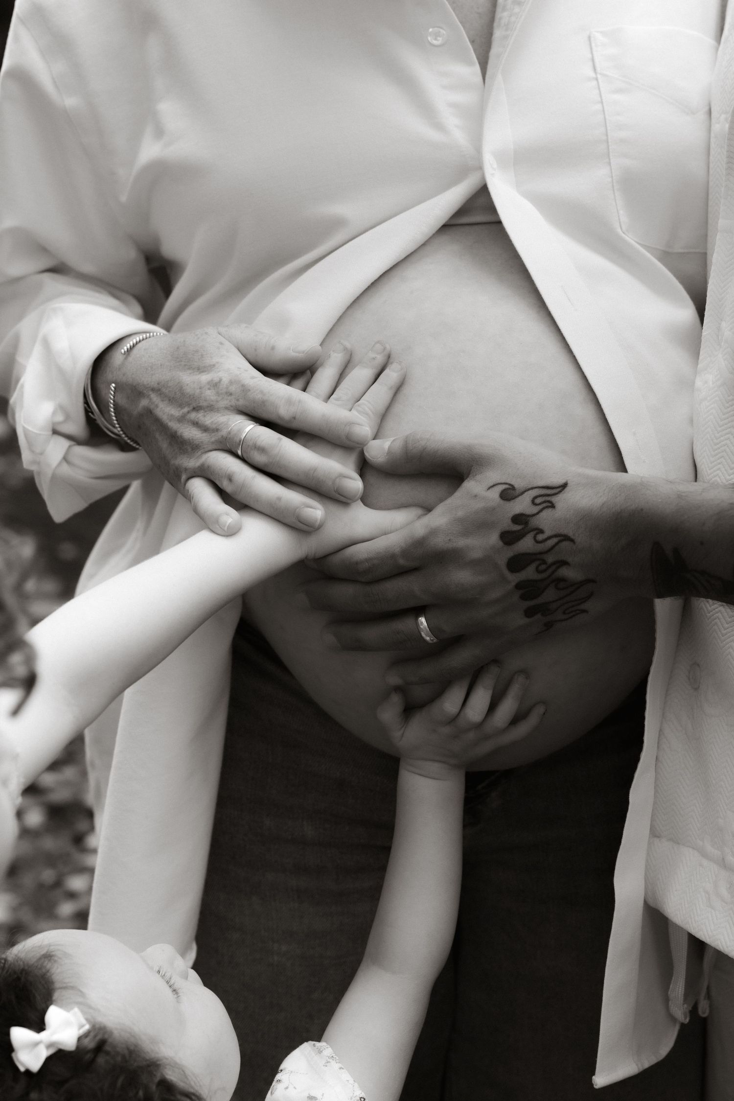 Black and white maternity photo showing multiple hands cradling a pregnant belly with artful tattoo detail.