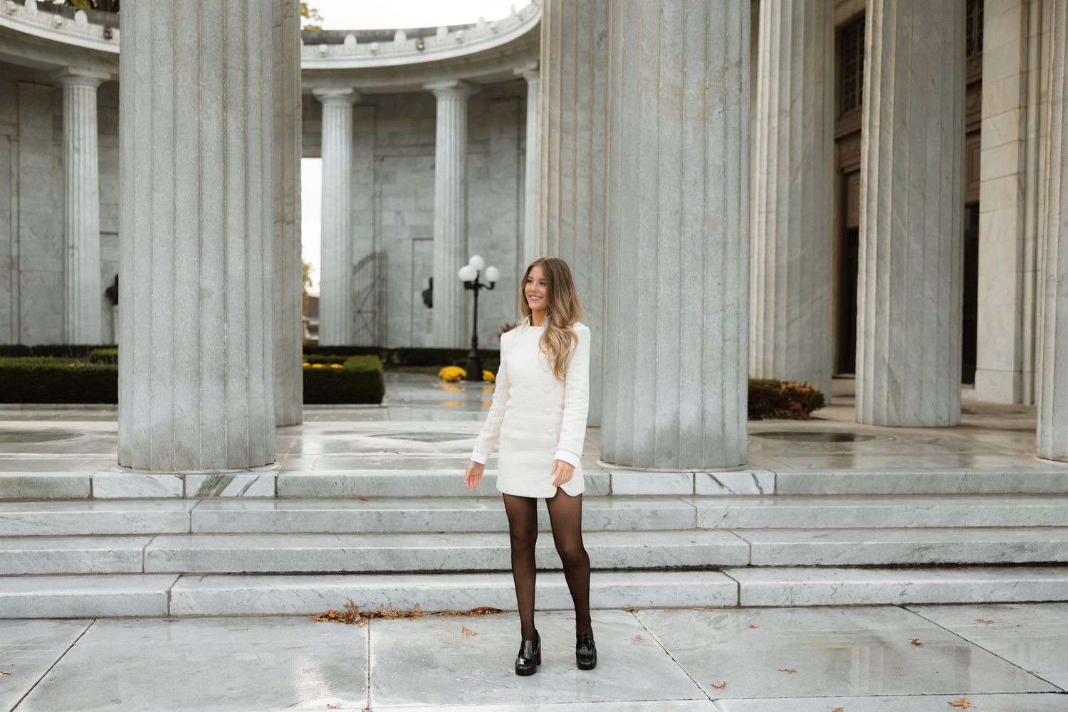 A person wearing a white blazer and black boots stands between classical stone columns in an architectural setting.