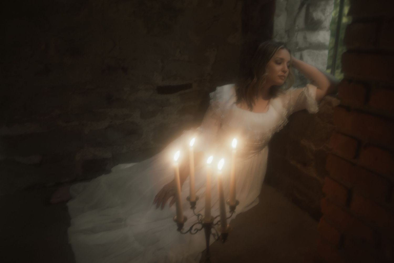 Atmospheric photo of person in flowing white dress sitting beside glowing candelabra in dark moody setting.