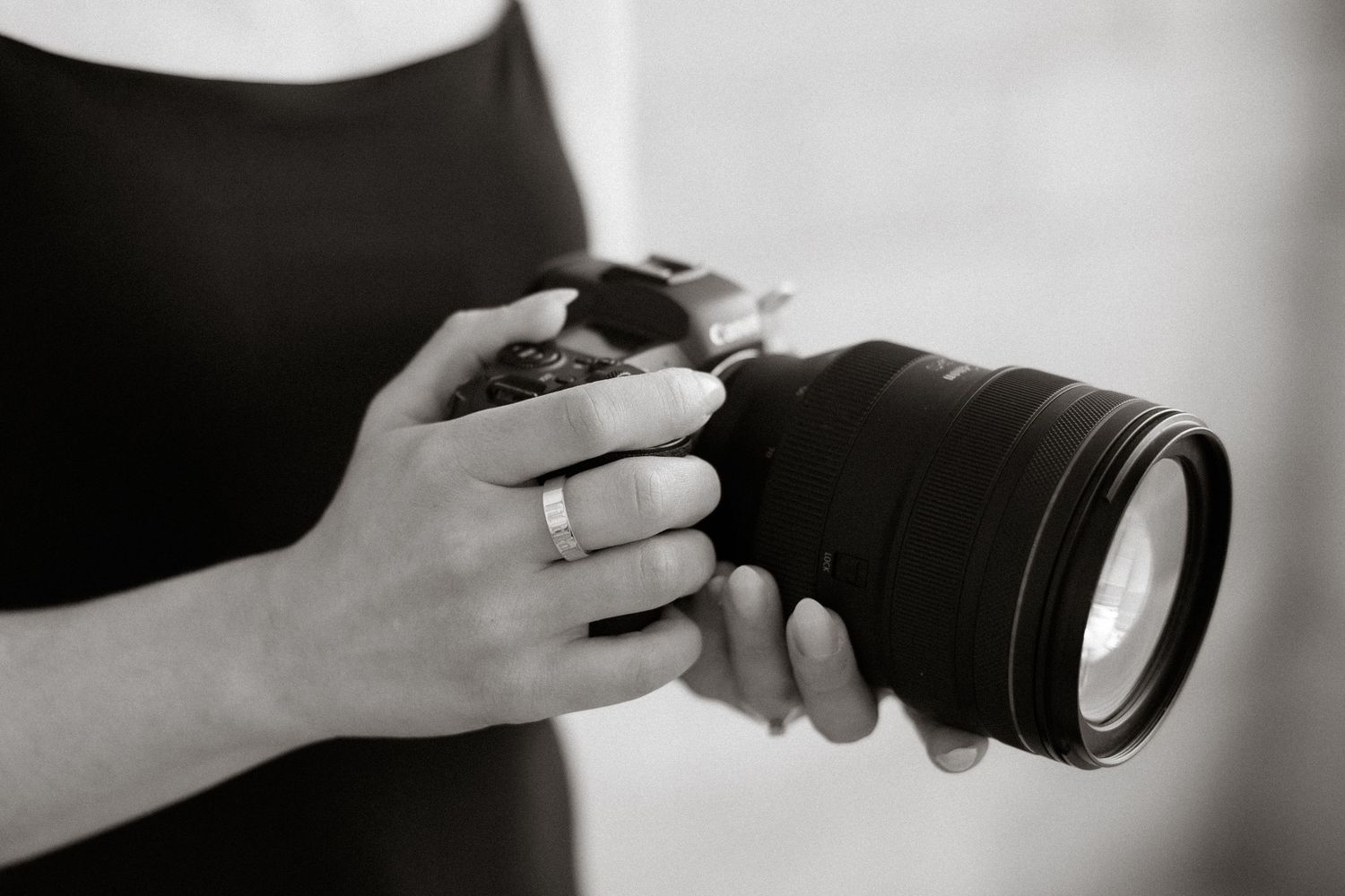 Black and white photo of a woman holding a DSLR camera with a large lens, wearing a ring on her finger.