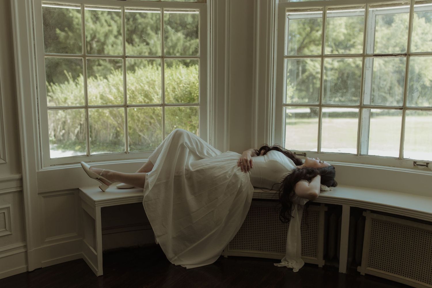 Bride to be draped over a sunlit window seat in an elegant bay window with tranquil garden view.
