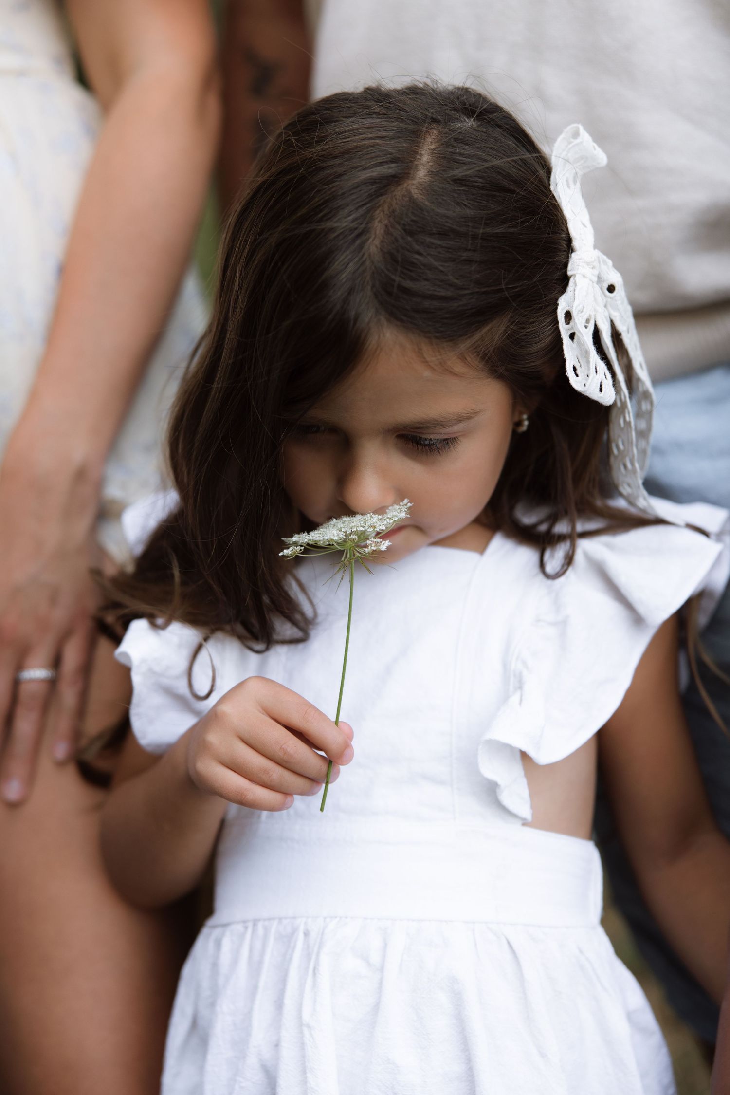 A young child wearing a white dress holds and examines a small white flower.