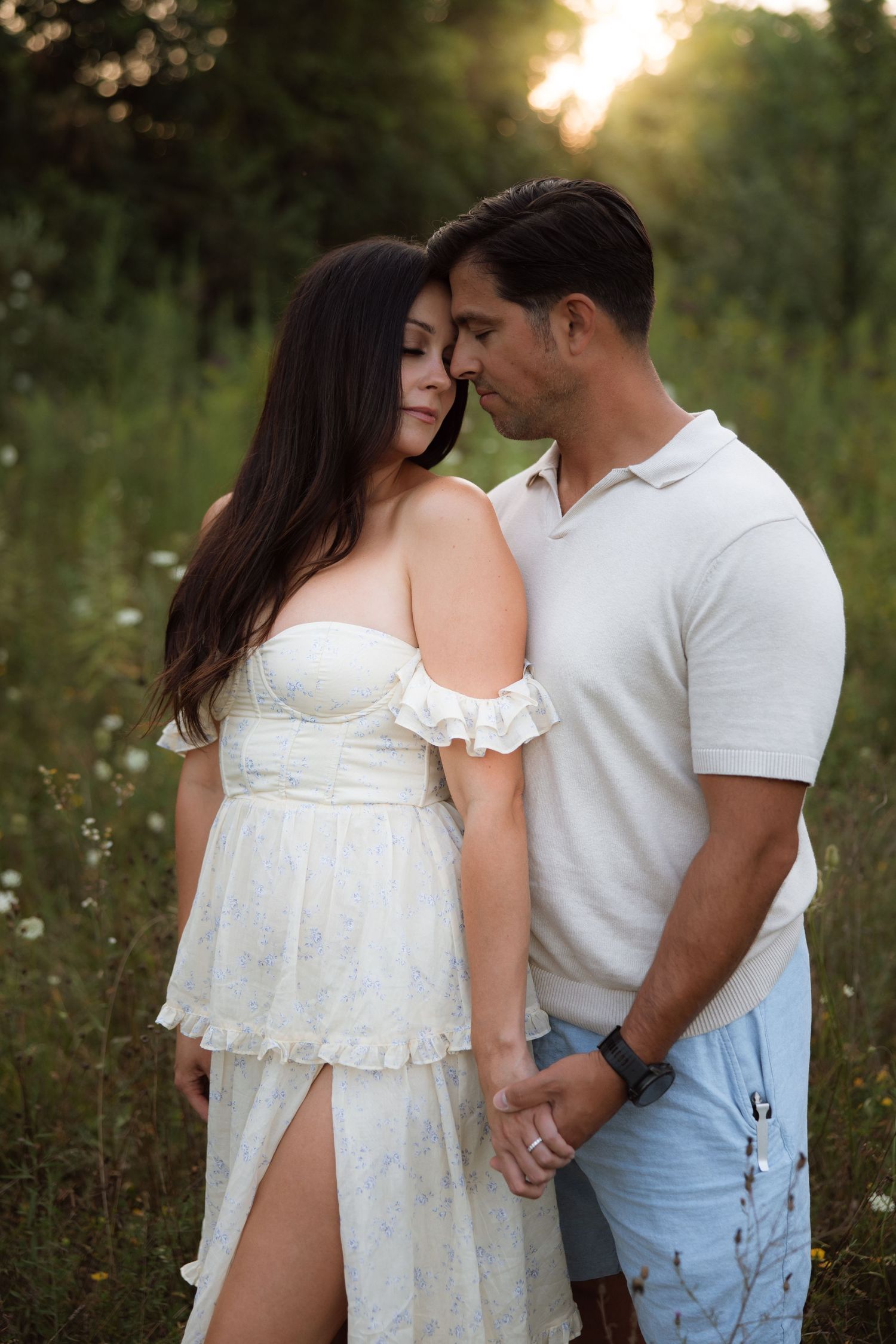 A couple in casual white outfits share an intimate moment together in a sunlit summer meadow at sunset.
