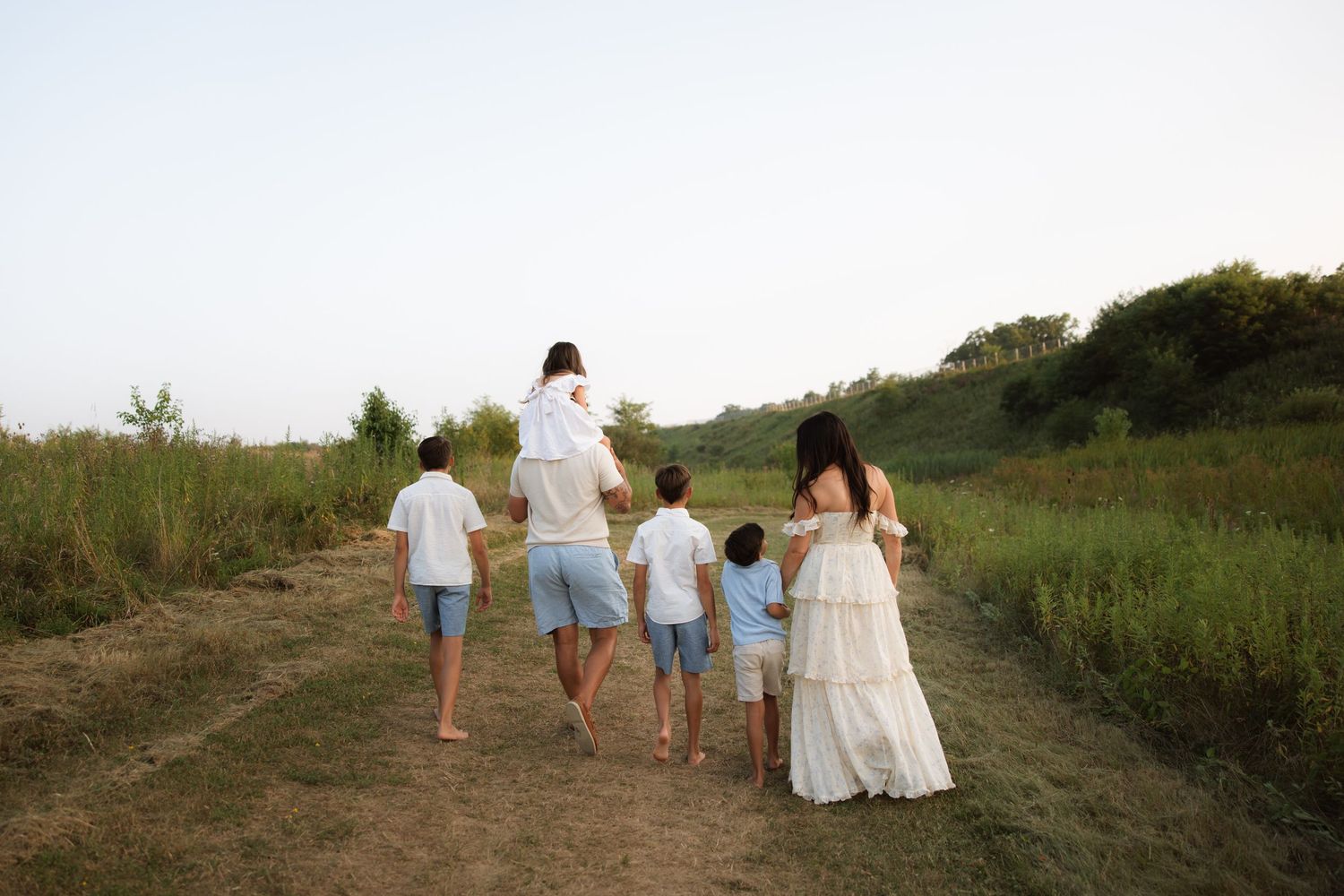 Wedding party wearing white walks through a coastal field at sunset, creating silhouettes against the golden sky.