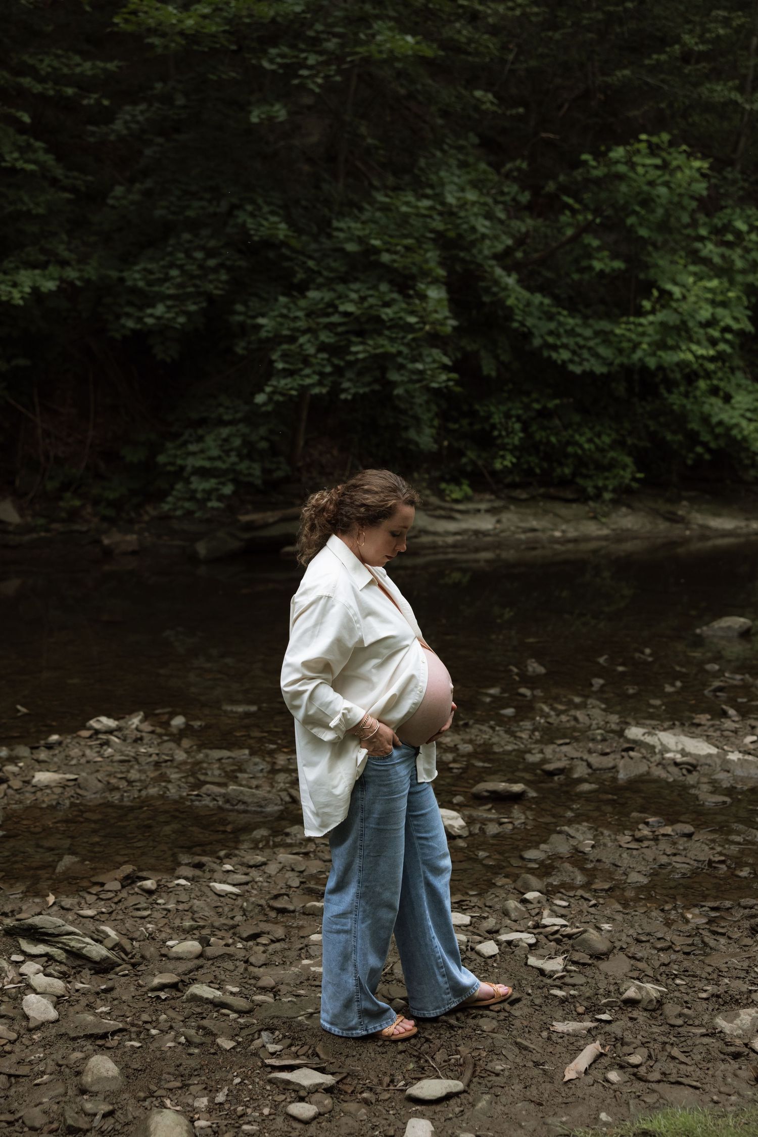 A pregnant person in a white shirt and jeans stands near a rocky riverbank surrounded by green trees.