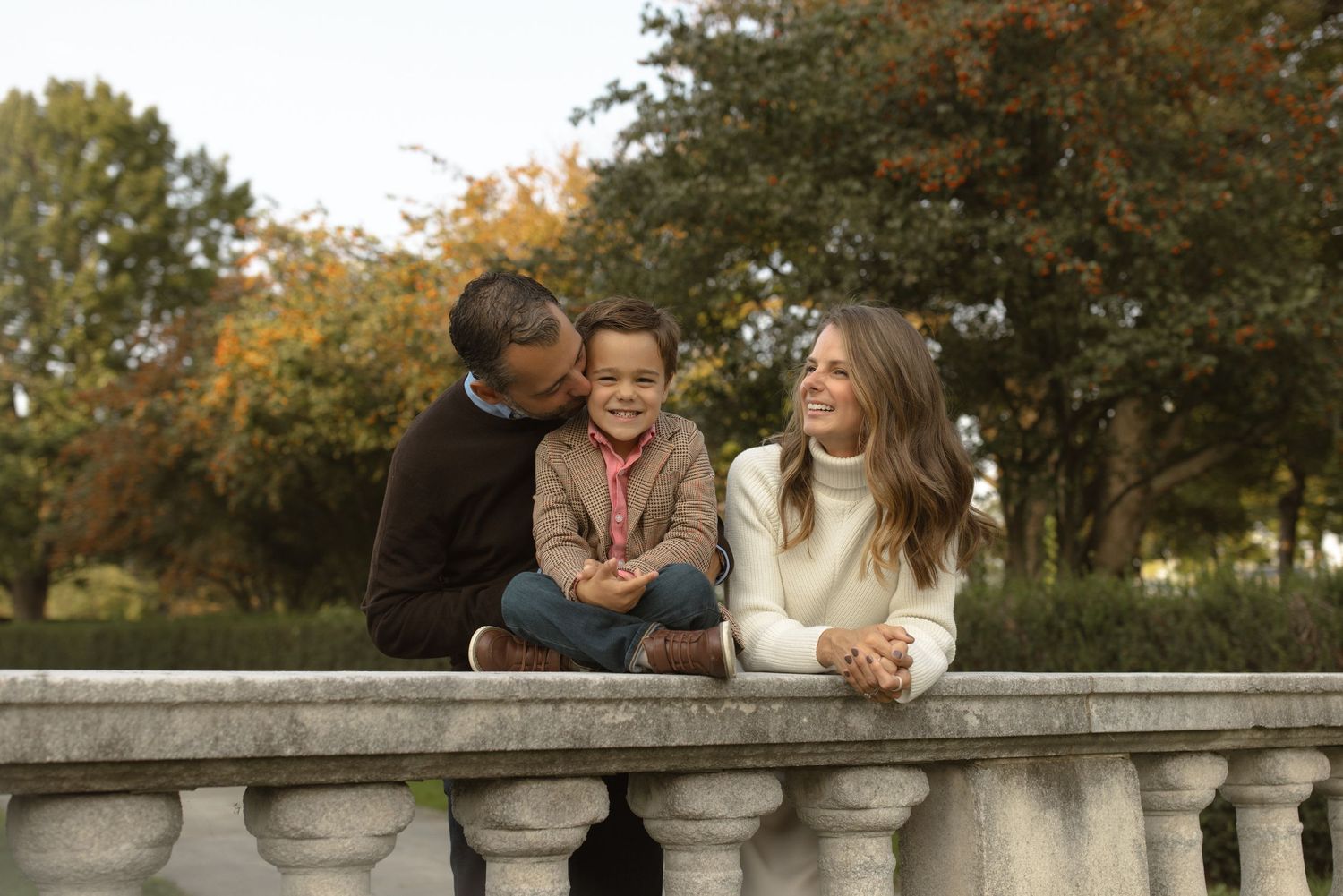 Three people share a joyful moment together while sitting on a stone balustrade during autumn.