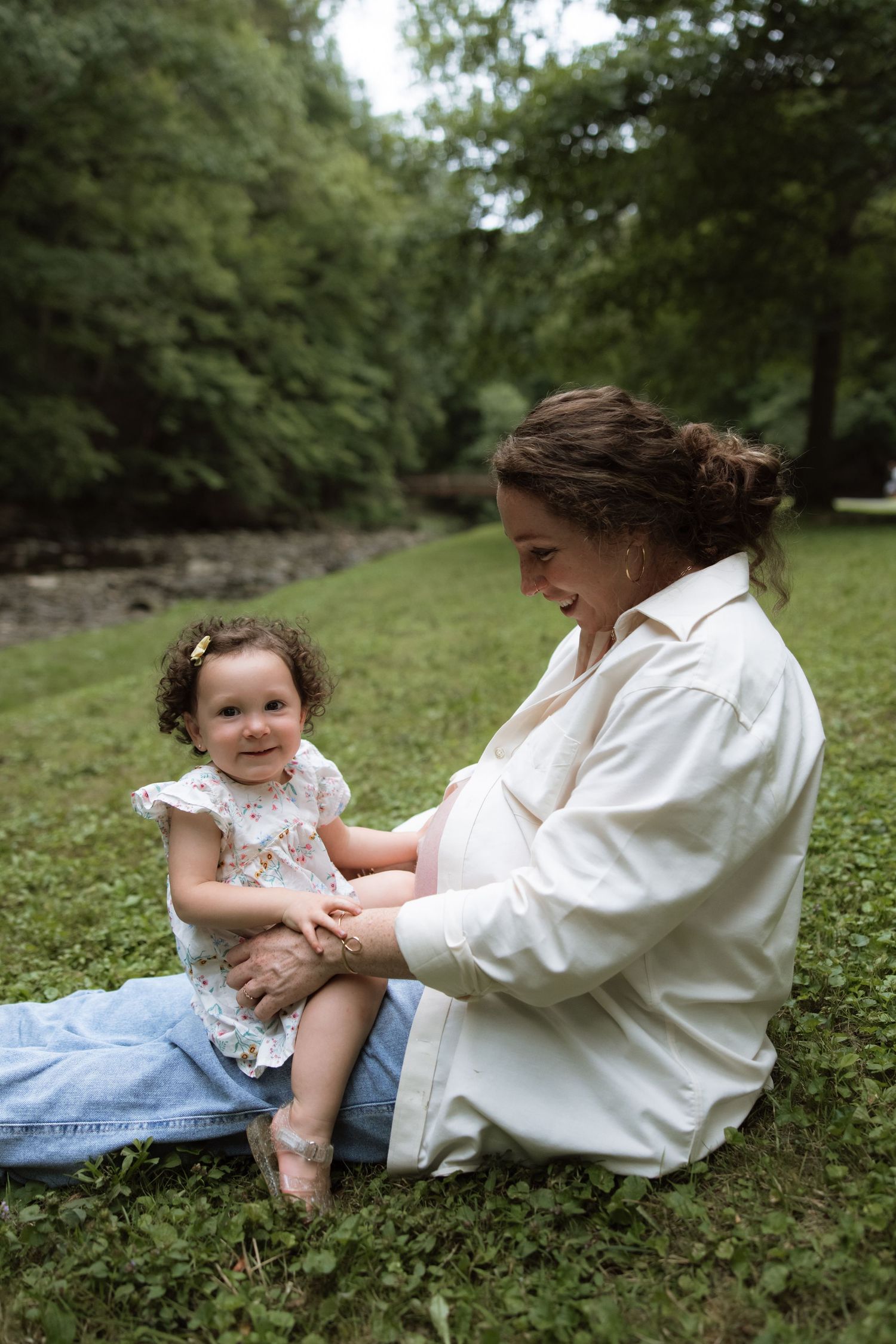 A touching moment between a parent and young child sitting on grass by a forest stream.