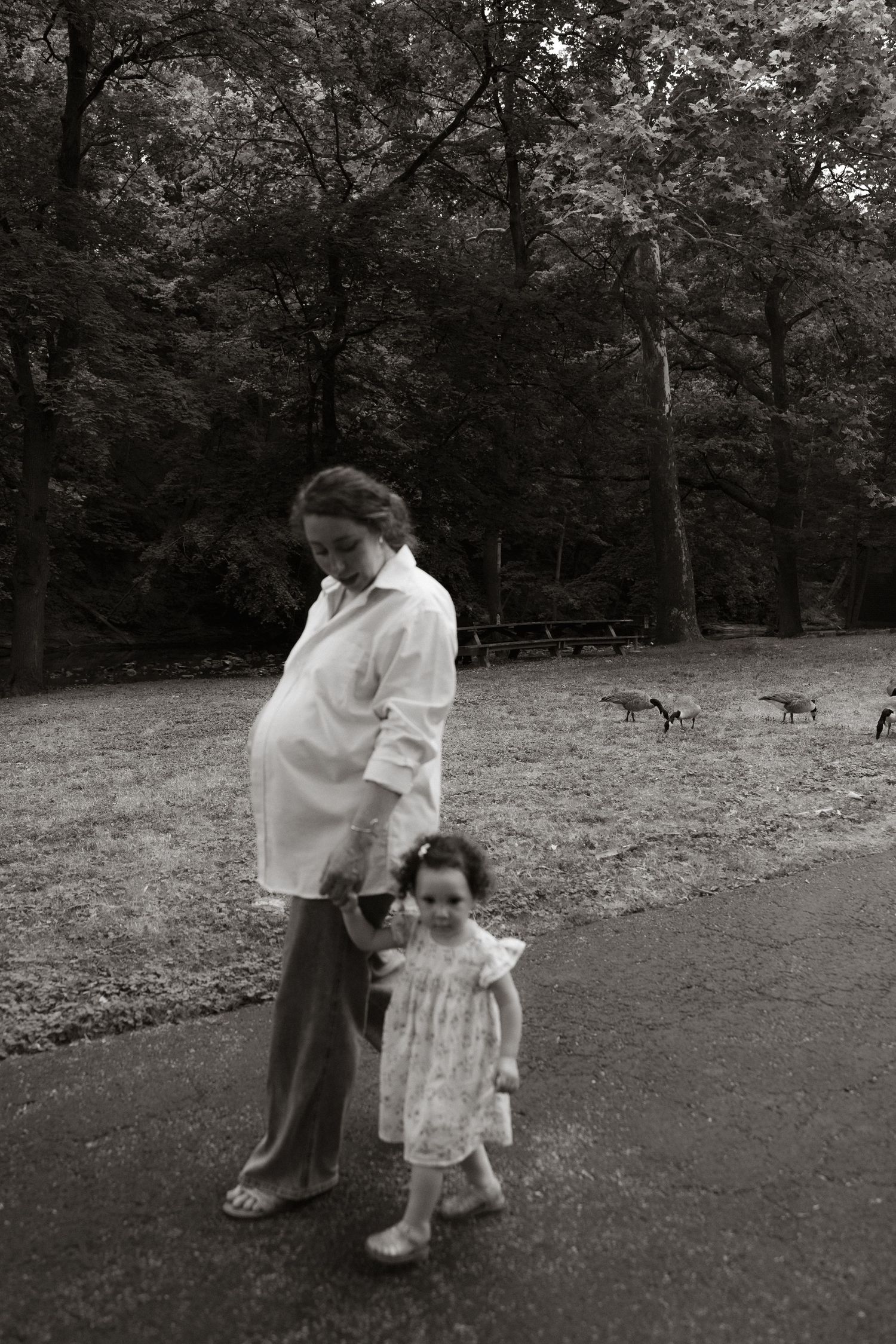A black and white photograph of a pregnant woman and small child walking outdoors in a park with trees.