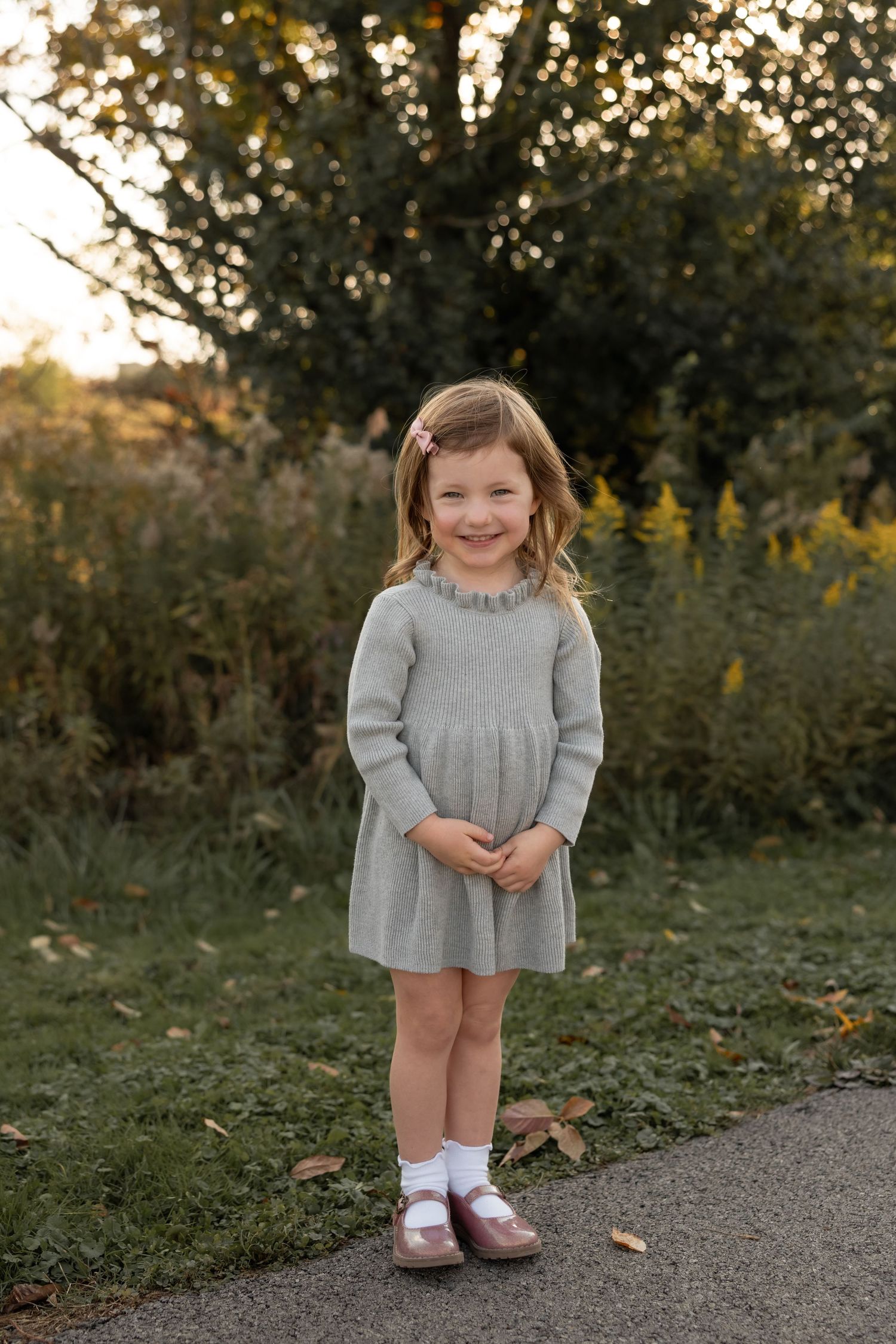 Child wearing a cozy gray sweater dress and pink shoes stands on a path during golden hour with trees in background.