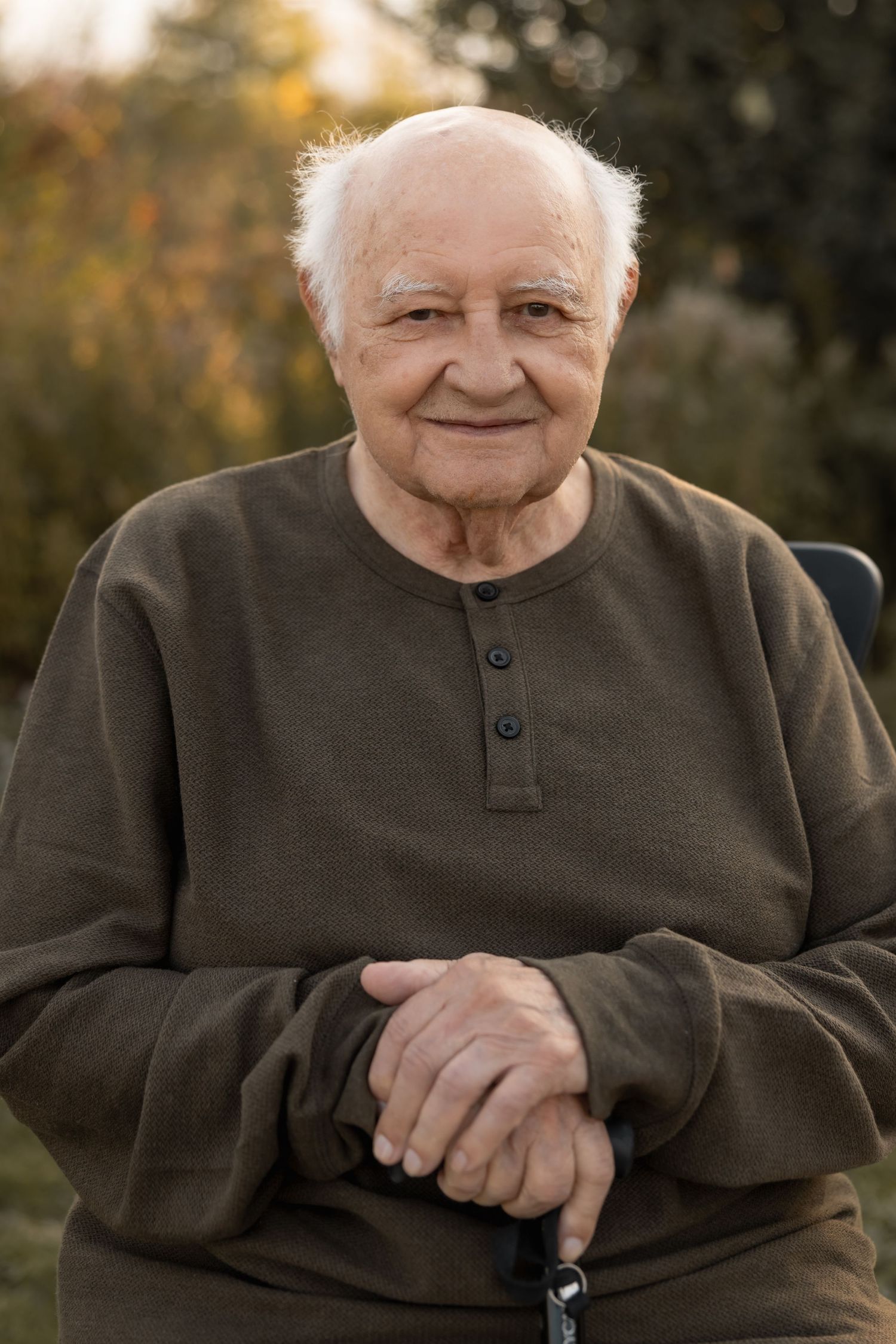 Elderly person wearing brown henley sweater sitting outdoors with clasped hands and warm smile on autumn day.