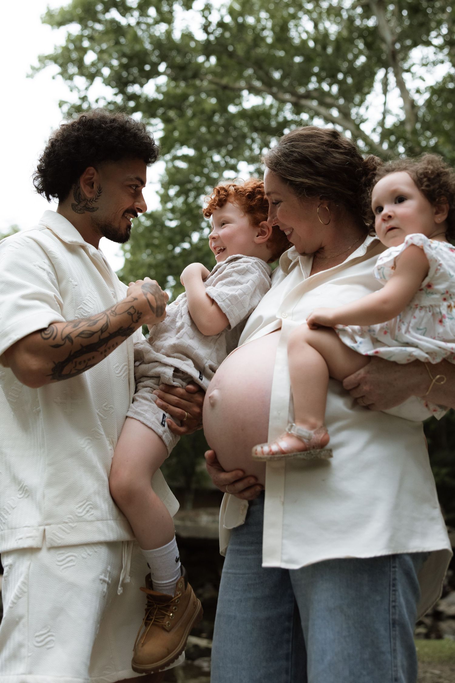 Family in white outfits sharing tender moment outdoors with children and pregnant belly.