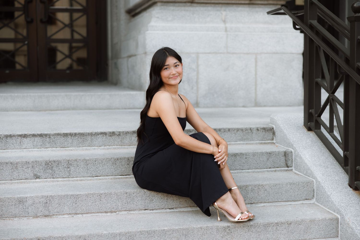 A figure in a black dress sits gracefully on white marble steps while posing for a photoshoot.