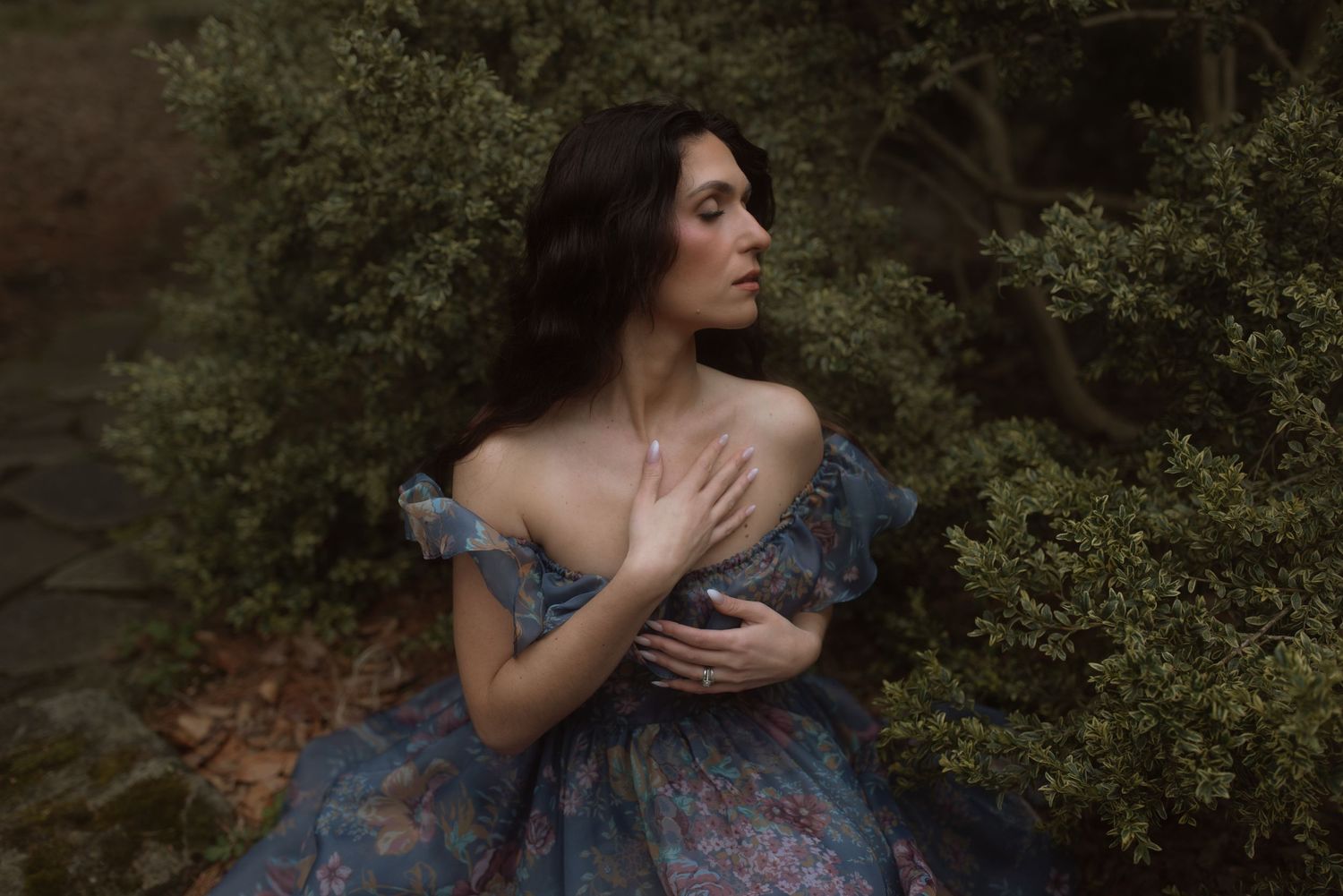 A woman in a blue vintage dress poses dramatically against lush greenery in soft lighting.