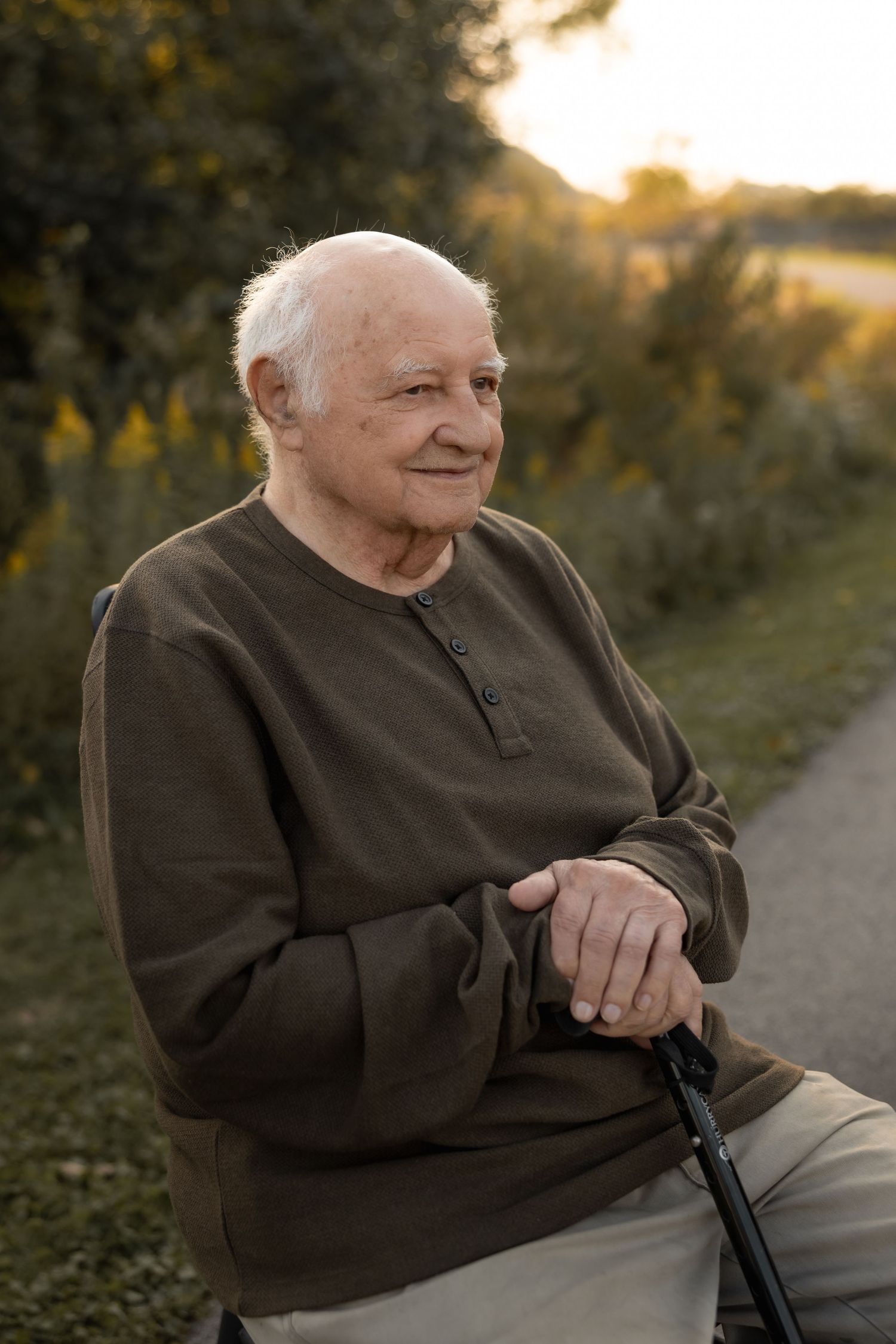 Elderly person in brown sweater sitting outdoors at sunset smiling joyfully during a golden hour photo session.