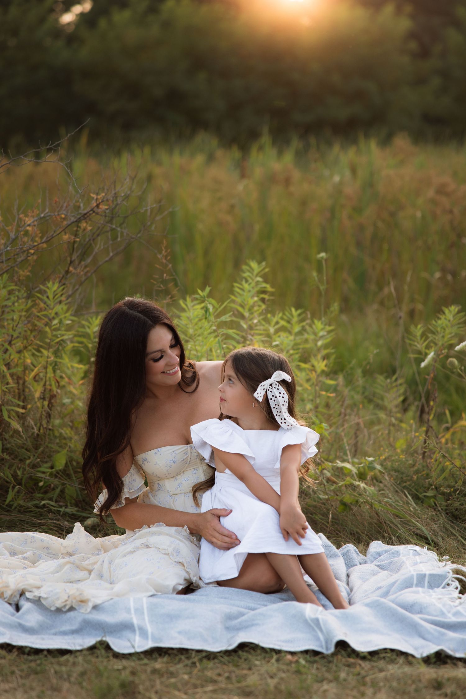 A mother and daughter share a tender moment lying on a white blanket in a sunlit meadow with tall grass at sunset.