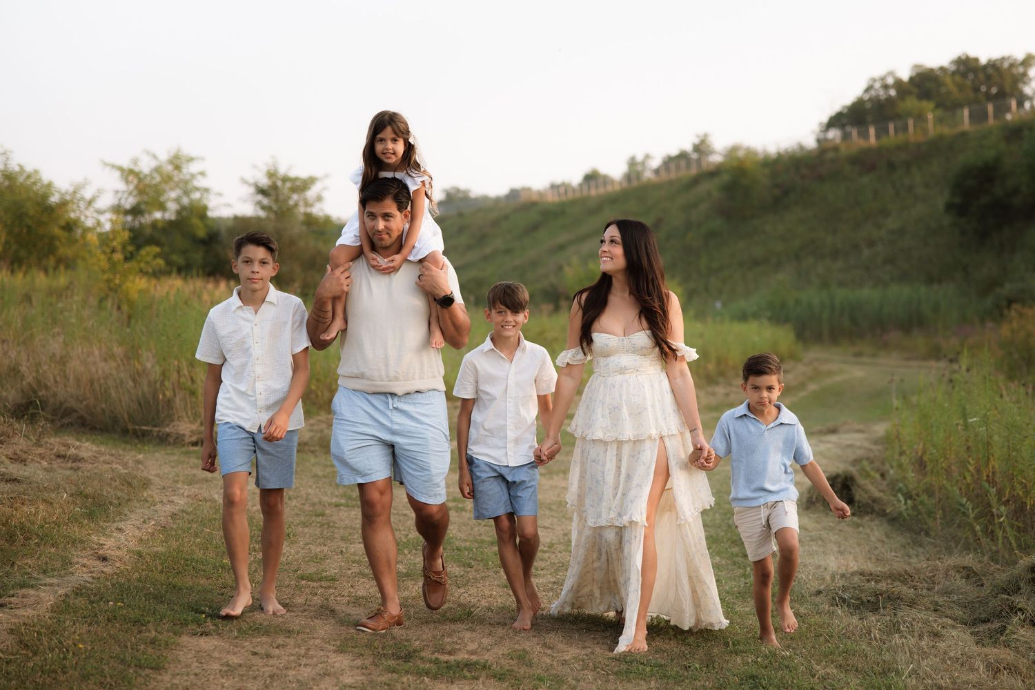 A family of five walks together barefoot on a grassy hill during sunset, wearing coordinating white and blue outfits.