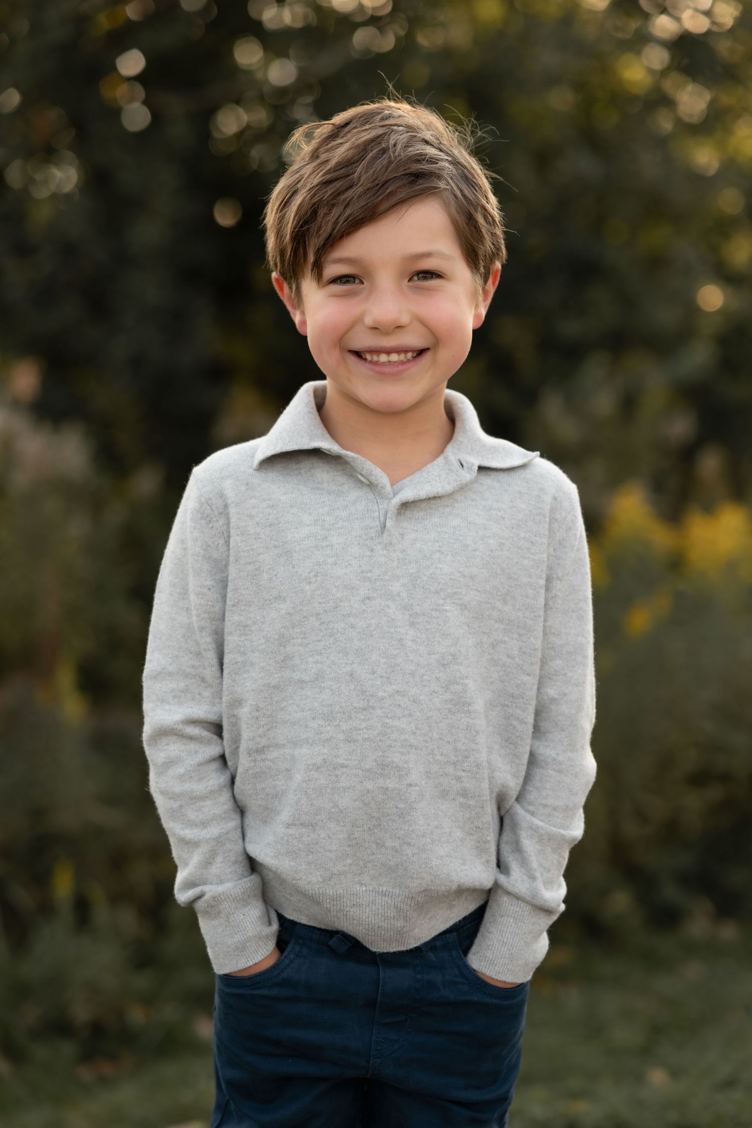 Child wearing a gray sweater stands outdoors with trees and bokeh lighting in the background during golden hour.