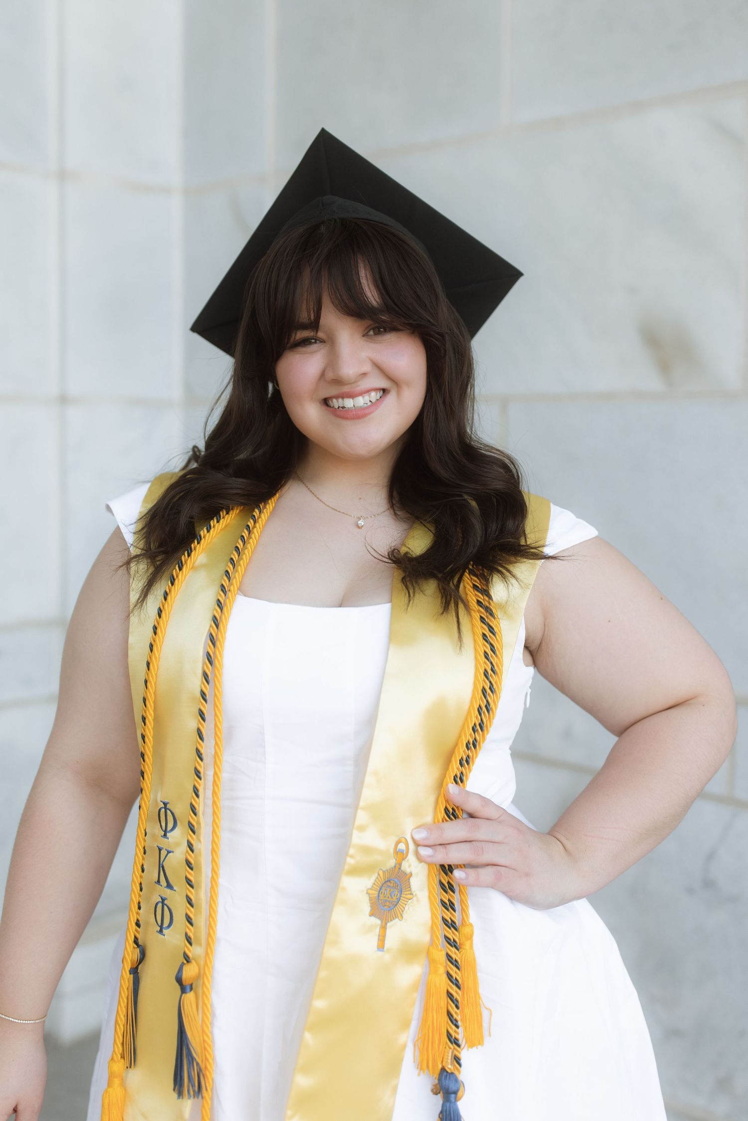 Graduate in white dress and gold sash wearing mortarboard cap poses for graduation portrait with hand on hip.
