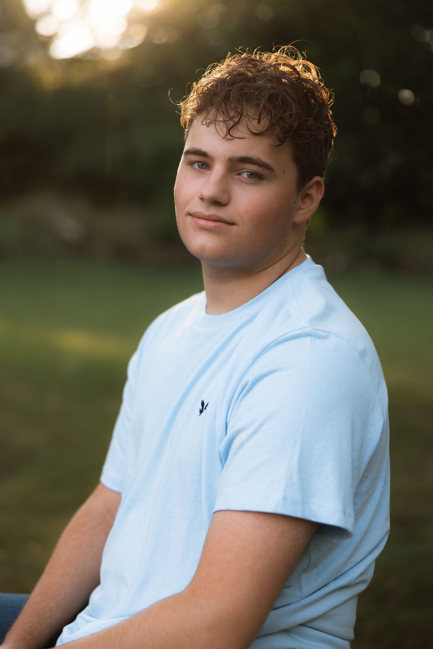 A person in a light blue t-shirt sits outdoors during golden hour with a natural blurred background.
